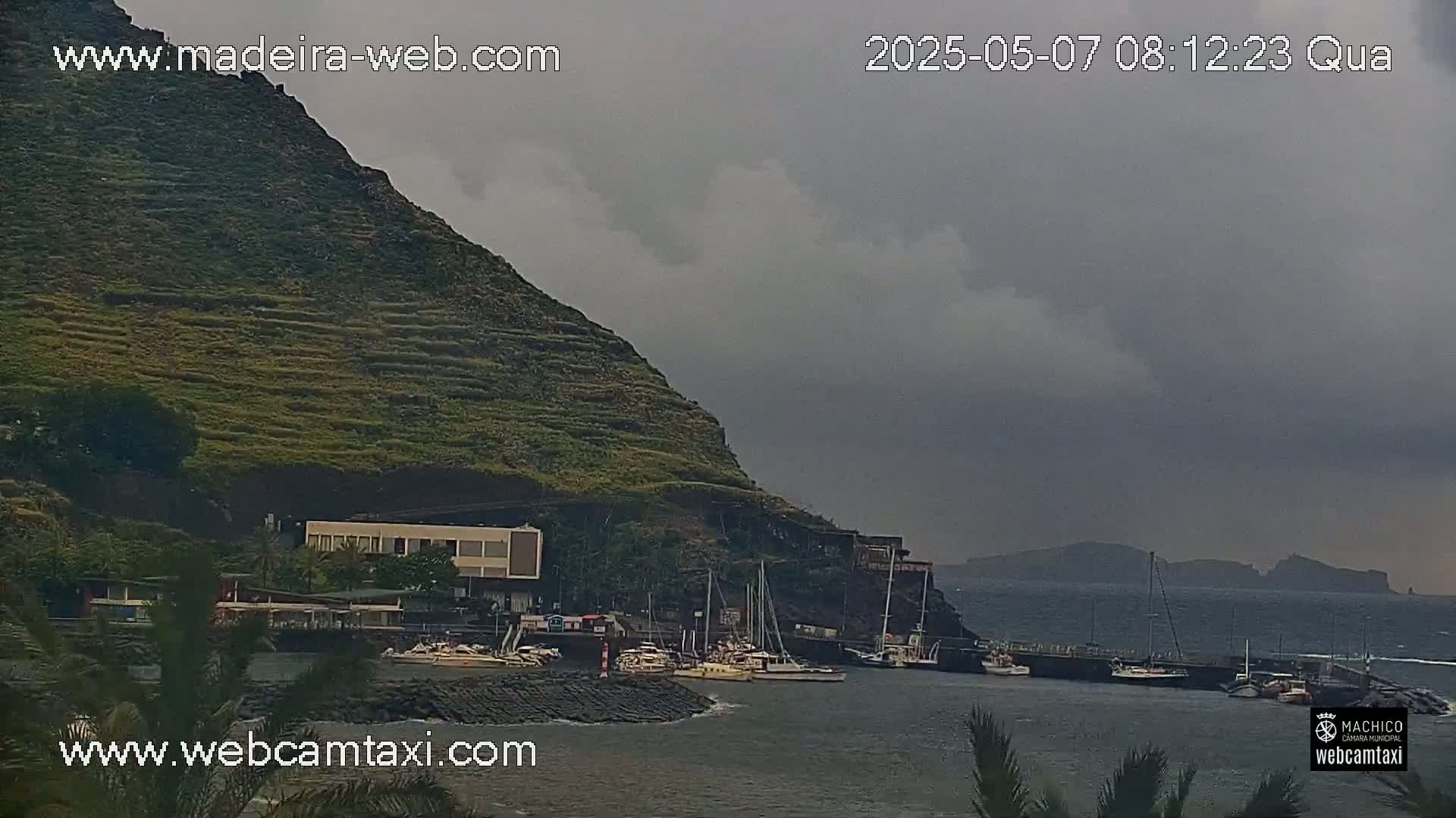 A harbor with several boats is nestled below a terraced hillside under a cloudy, overcast sky.