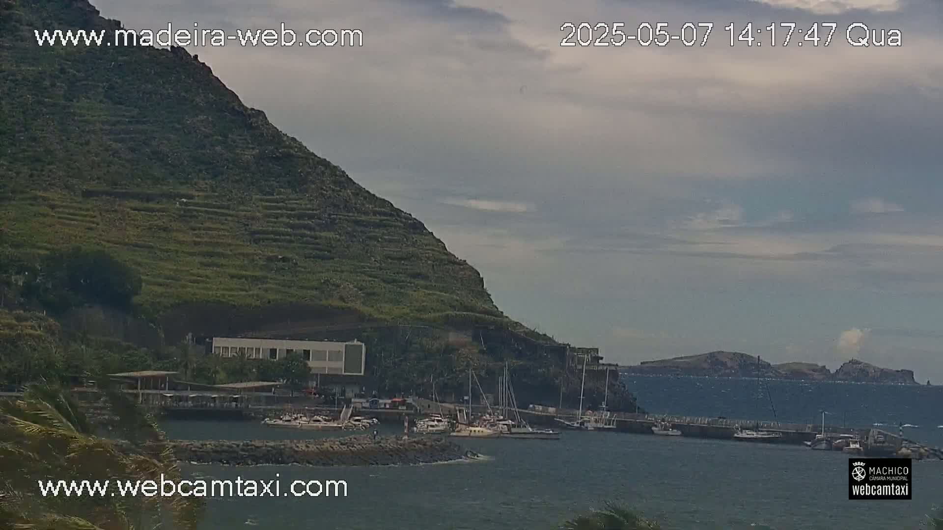 A small marina with boats is nestled below a terraced hillside on a partly cloudy day.