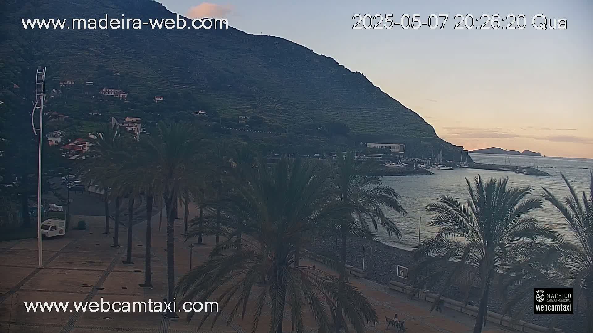 A coastal scene at twilight shows palm trees lining a paved area next to a calm bay with boats, a hillside village, and a dark mountain in the background.