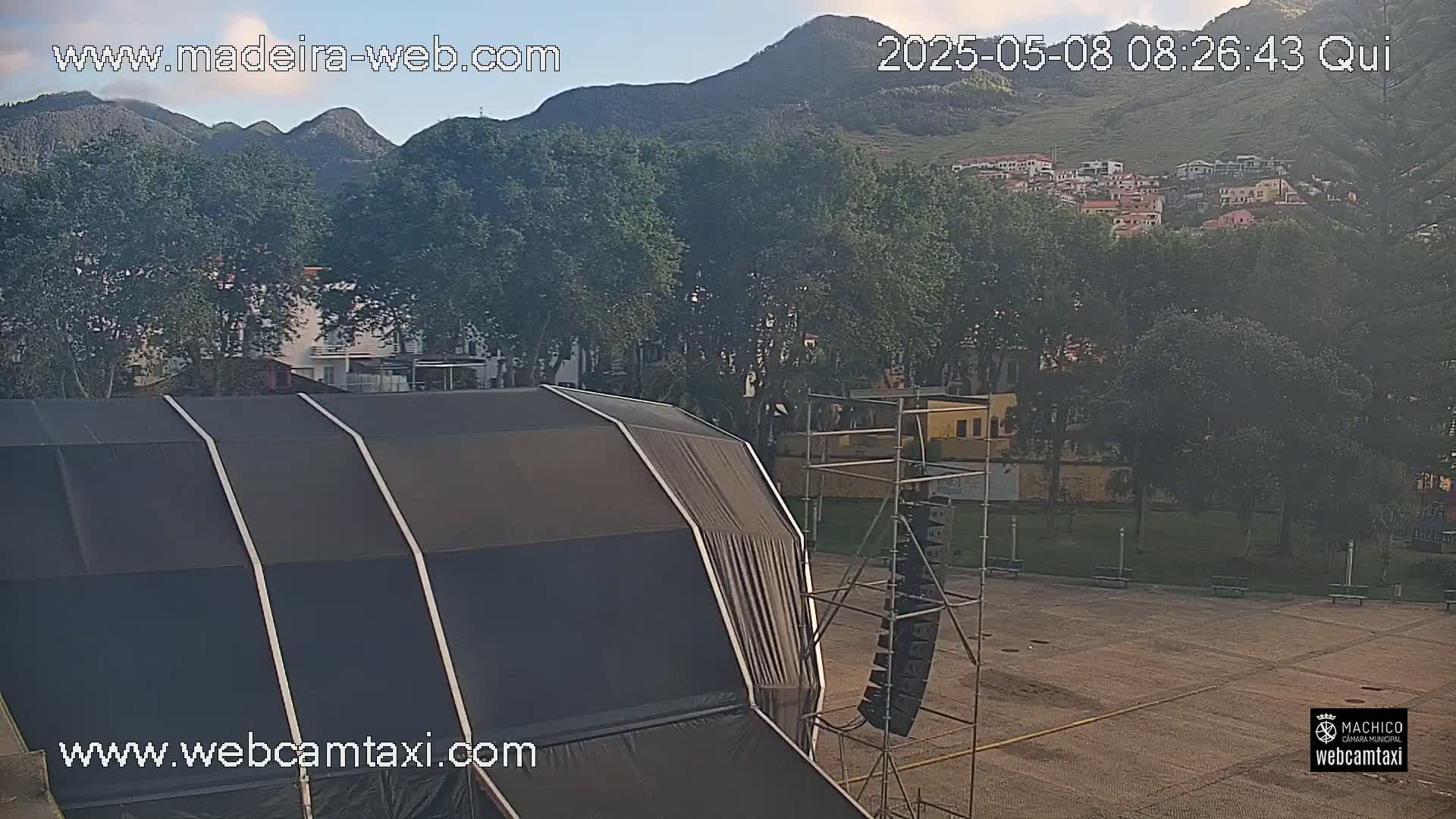 A large black tent sits in a paved area next to a scaffolding structure with speakers, all set against a backdrop of green trees, distant buildings, and a mountain under a mostly clear sky.