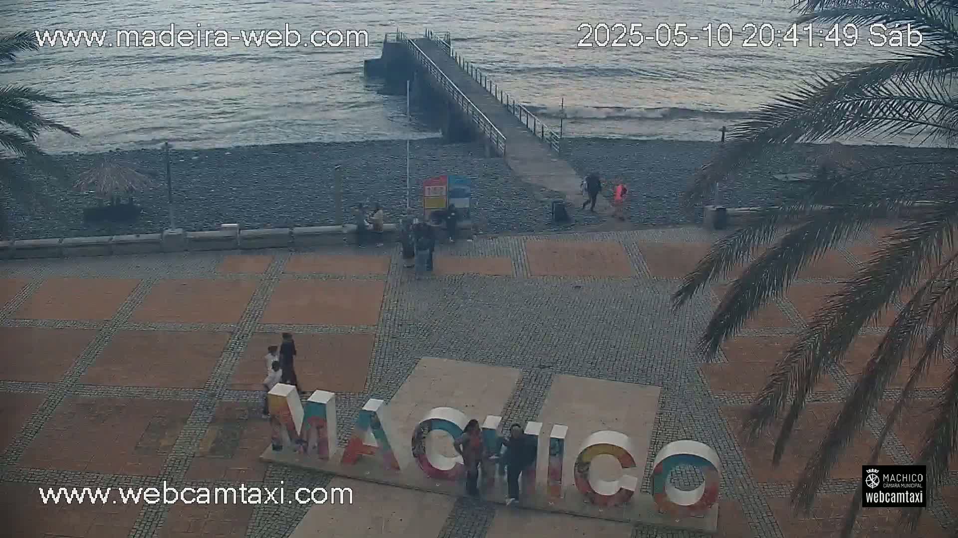 An overcast day shows a pebble beach with a pier, several people walking near a large sign that reads "Machico," and palm trees in the foreground.