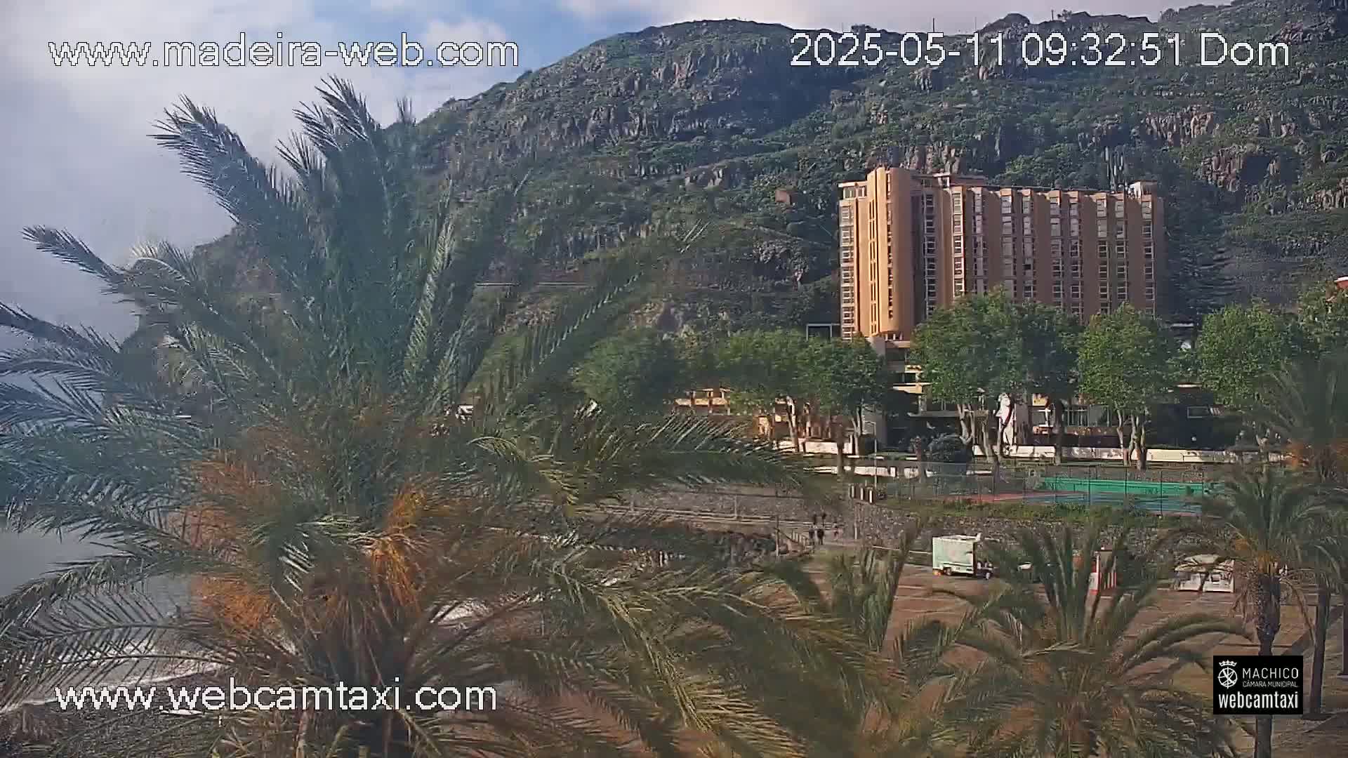A partly cloudy day shows a large hotel building situated near a rocky mountainside, surrounded by palm trees and other vegetation.