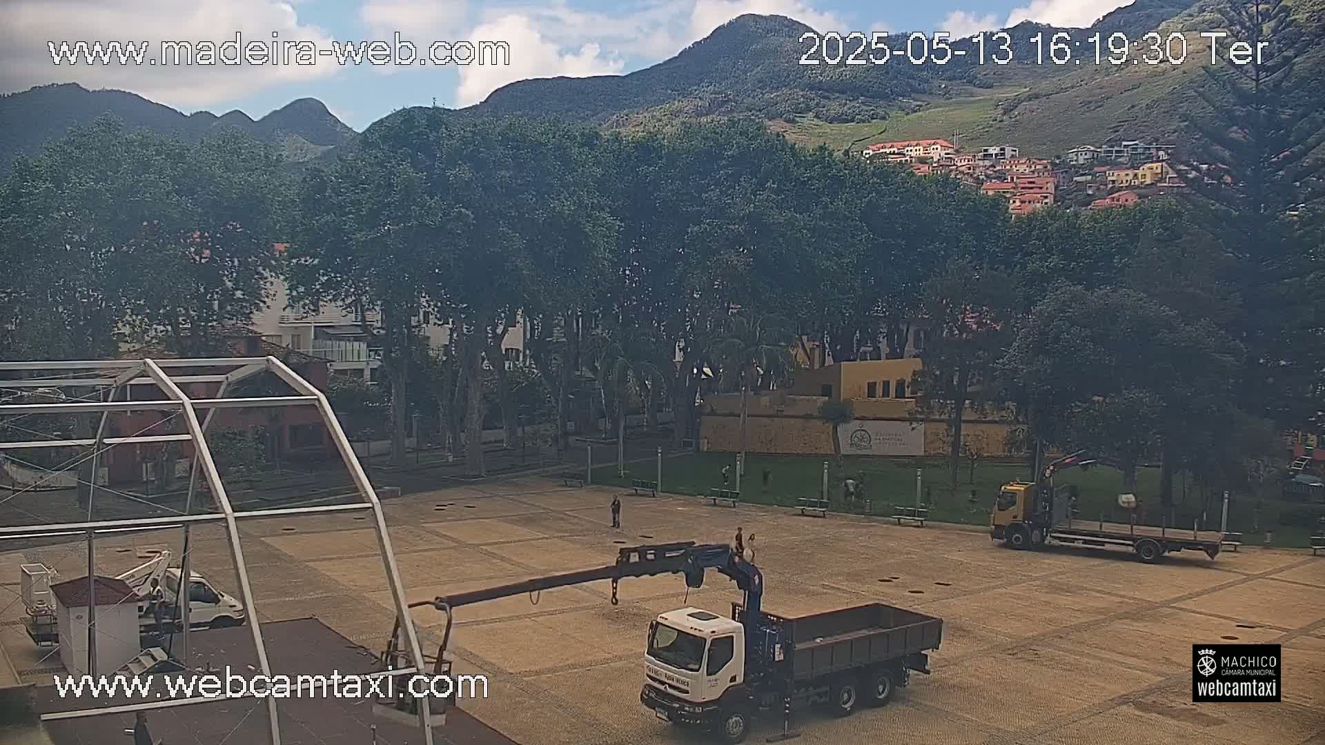 A partly cloudy day reveals a town square with several construction vehicles and a crane truck, backed by lush greenery and mountains.