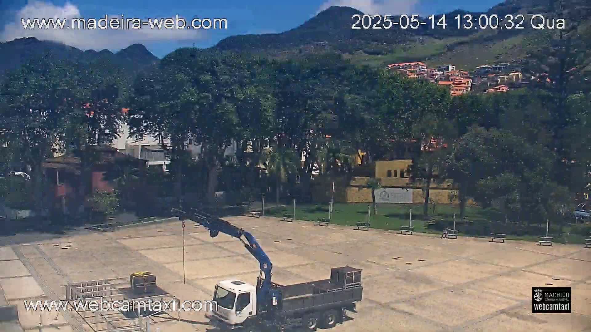 A crane truck is parked in a paved plaza, surrounded by lush green trees and buildings under a clear, sunny sky, with mountains visible in the background.