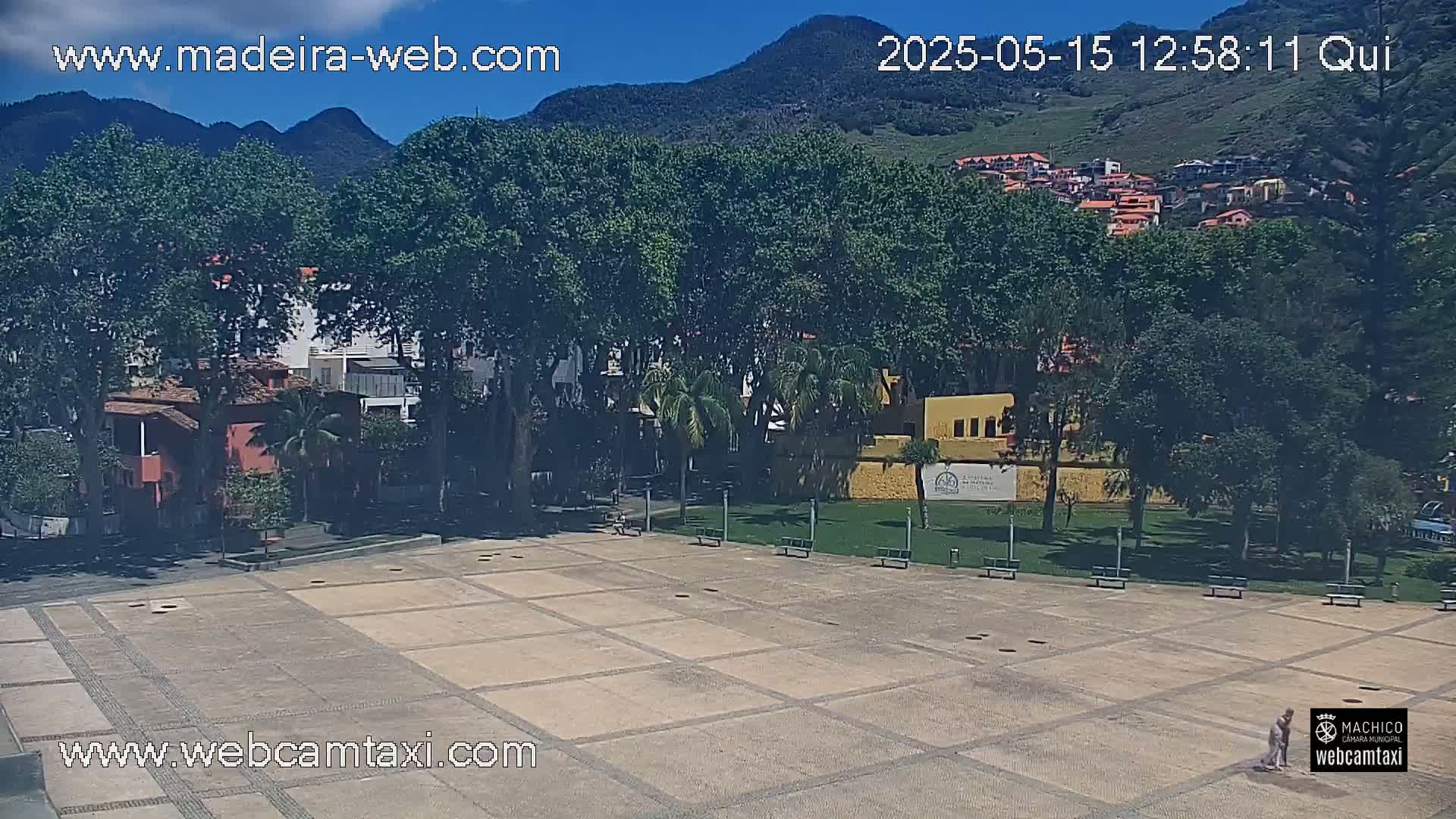 A paved plaza is surrounded by lush green trees and buildings, with mountains visible in the background under a clear, sunny sky.