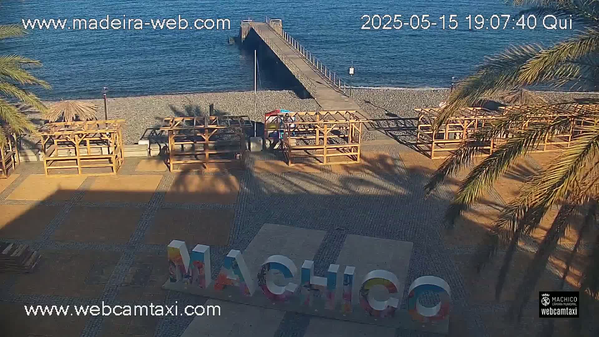 A beach with a pier, several empty wooden structures, and a large sign spelling out "Machico" under a sunny sky.