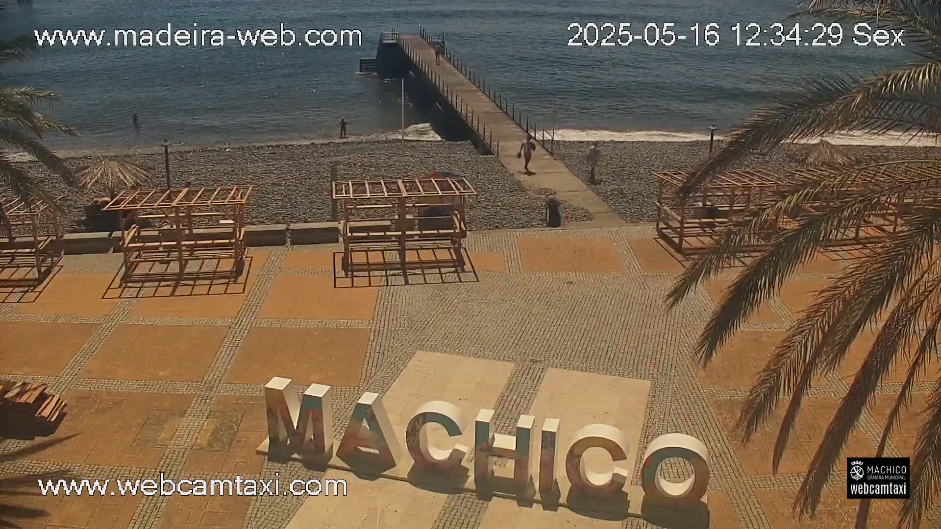 A sunny beach scene shows a paved area with several wooden structures, the word "Machico" in large letters, a pier extending into calm ocean water, and a few people walking along the shore.