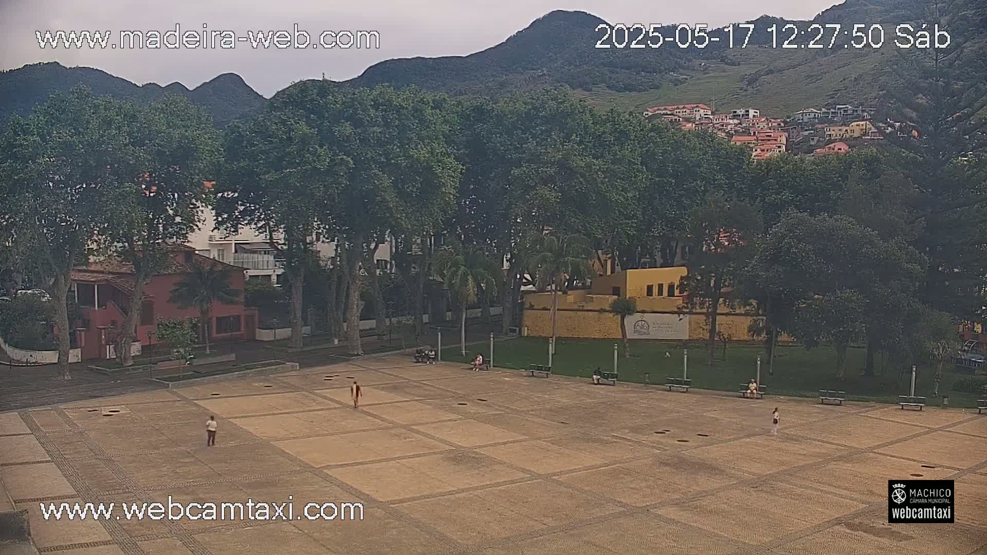 A paved plaza with a few people is surrounded by lush green trees and buildings, with mountains in the background under an overcast sky.