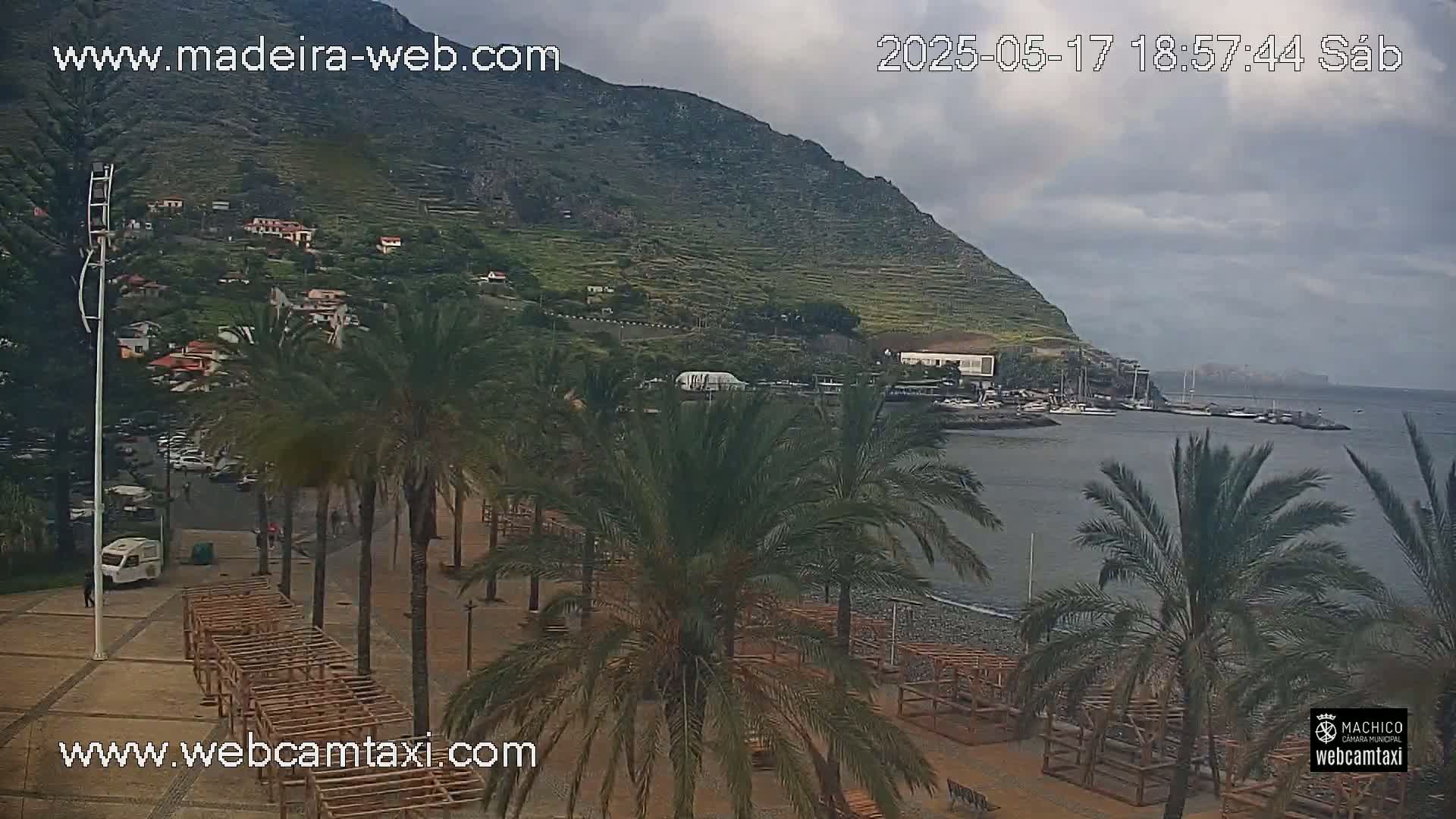 A coastal scene under a cloudy sky shows a pebble beach lined with palm trees and wooden structures, a calm body of water with boats, and a mountainous backdrop with buildings.