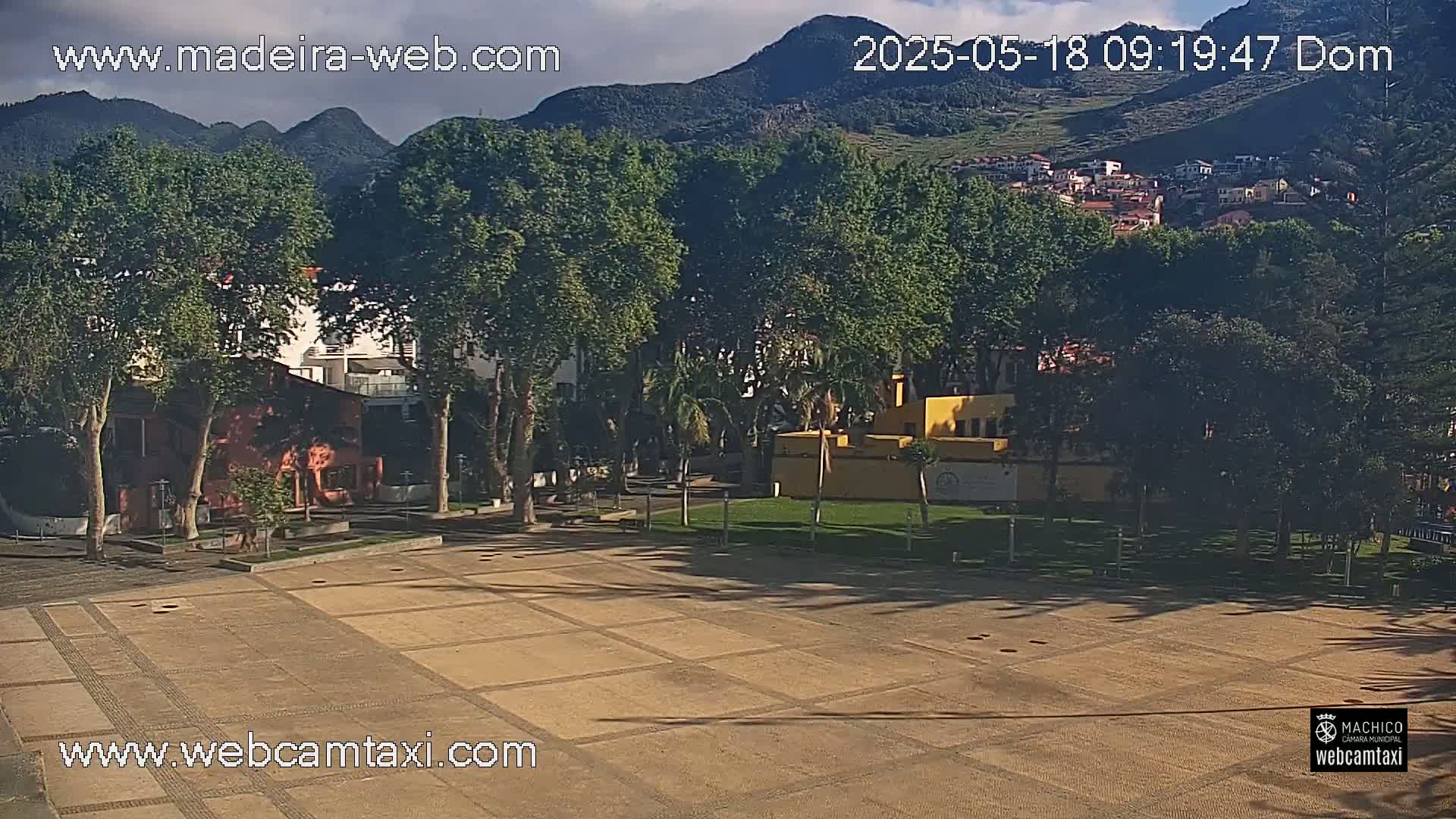 A paved plaza is visible, surrounded by trees and buildings, with mountains in the background under partly cloudy skies.