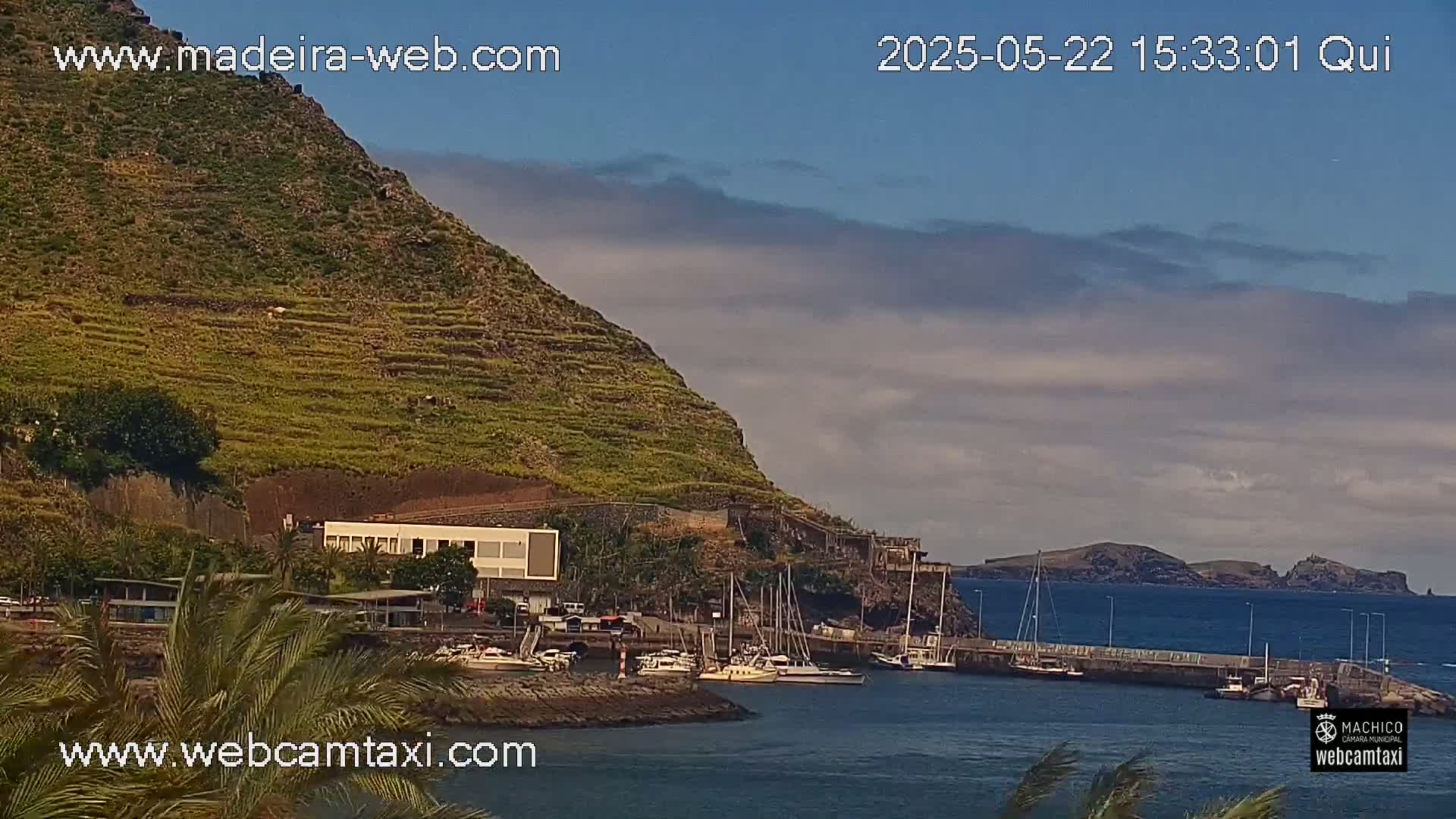 A small harbor filled with boats sits at the base of a terraced hillside under a partly cloudy sky.