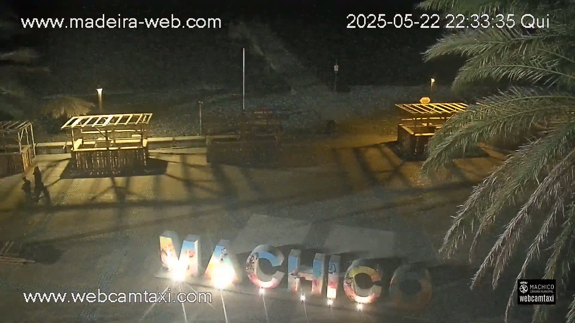 At night, an empty beach shows two wooden structures and a large illuminated sign, with palm trees visible in the foreground.