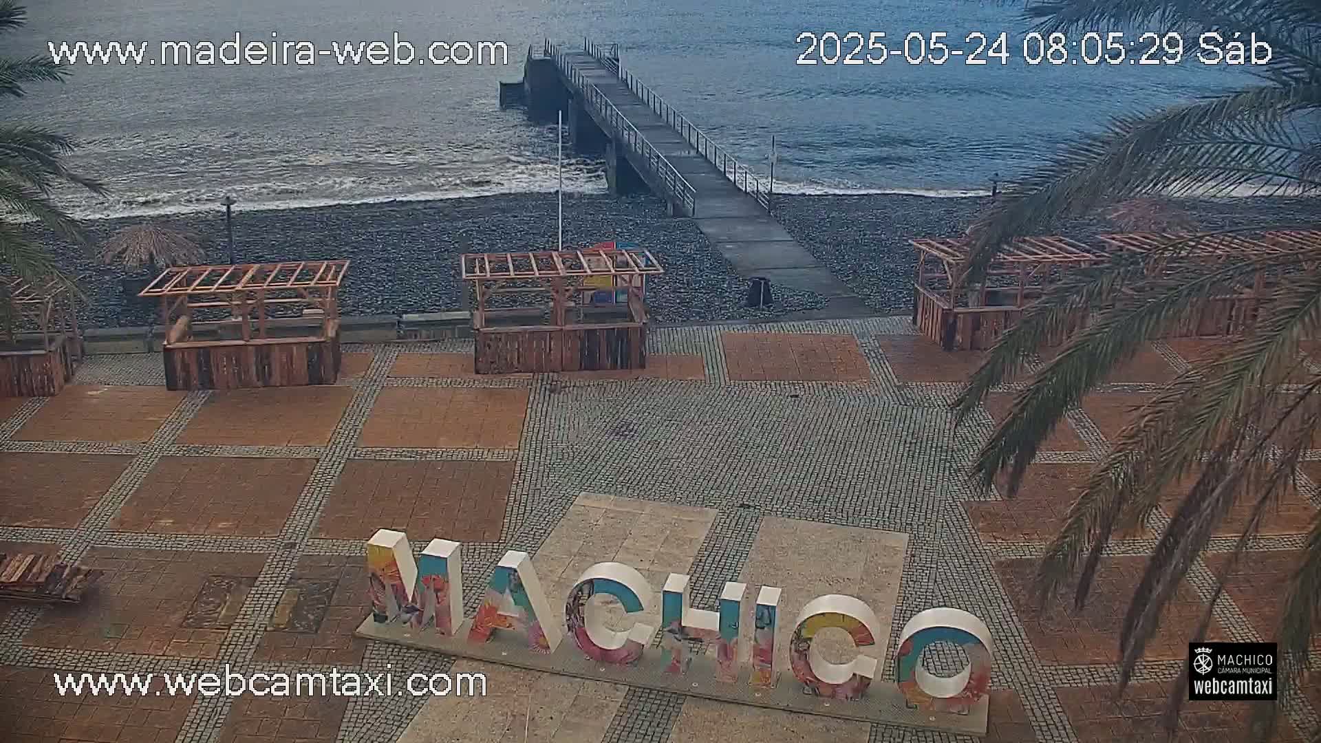 A paved plaza with several wooden structures and a large "Machico" sign is next to a pebble beach and a pier, overlooking a somewhat choppy ocean.