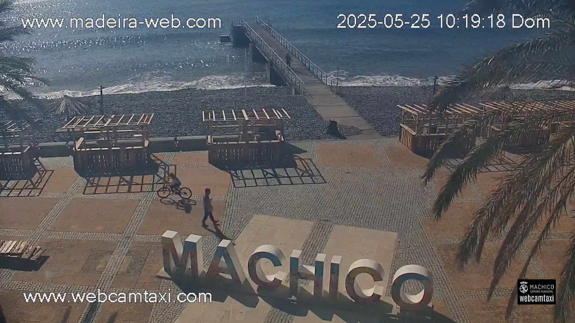 A paved beachfront area with several wooden structures, a pier extending into a calm ocean, a person walking, and a person riding a bicycle, all under sunny skies.