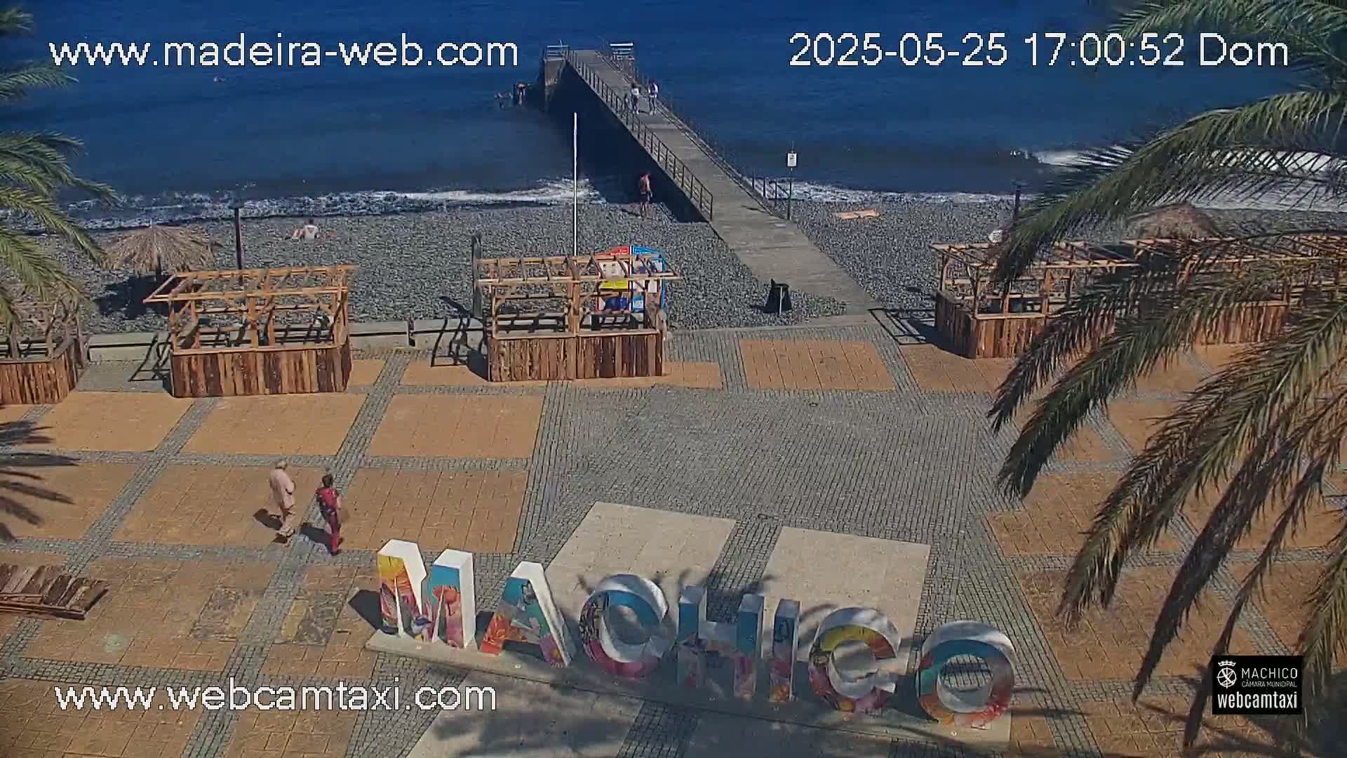 A beach scene with a pebble shore, wooden beach huts, a pier extending into a calm ocean, and a large "Machico" sign on a paved area, all under a sunny sky.