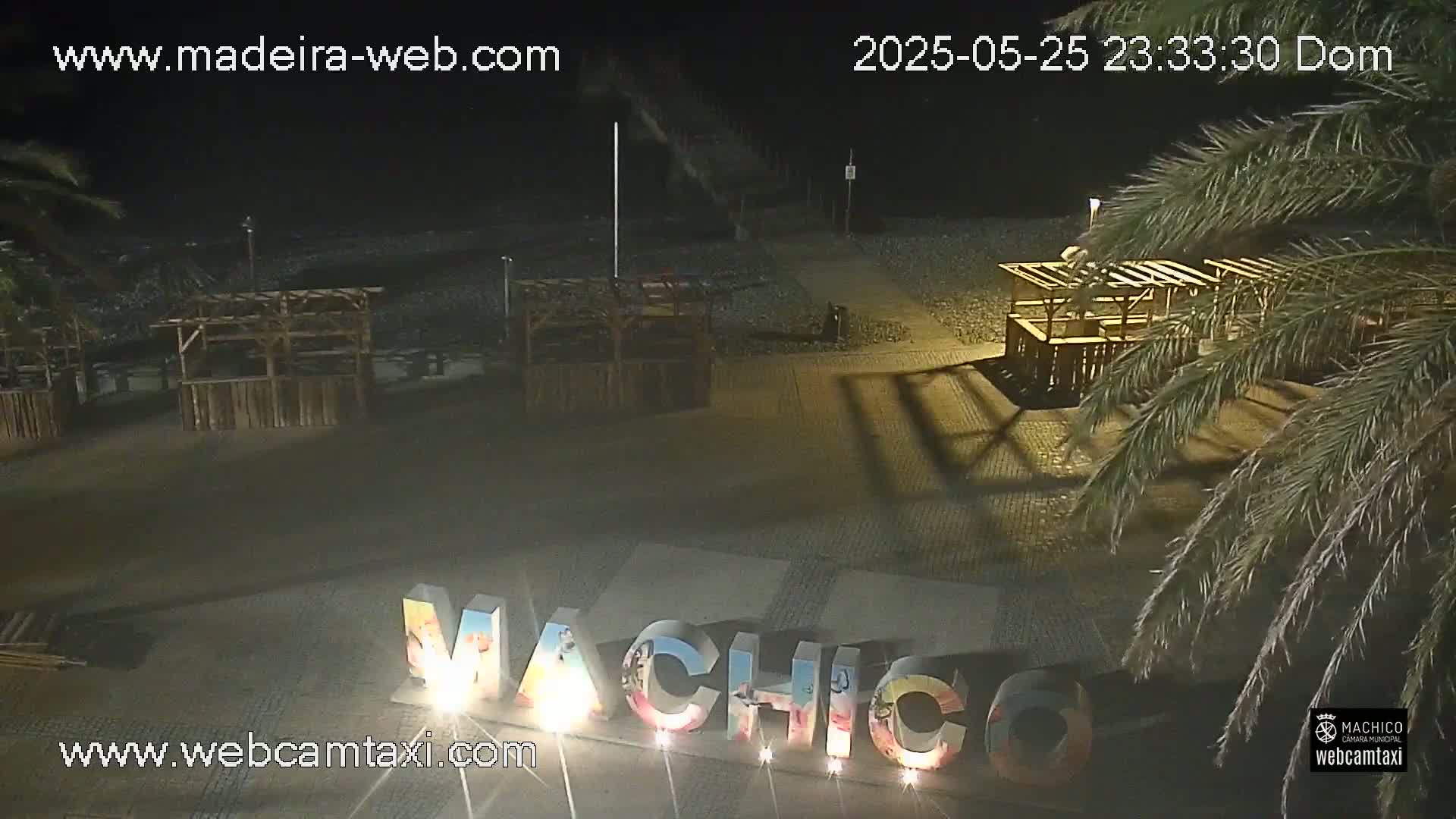 A nighttime view of a beach at night with several empty wooden structures, palm trees, and large illuminated letters spelling out a place name.