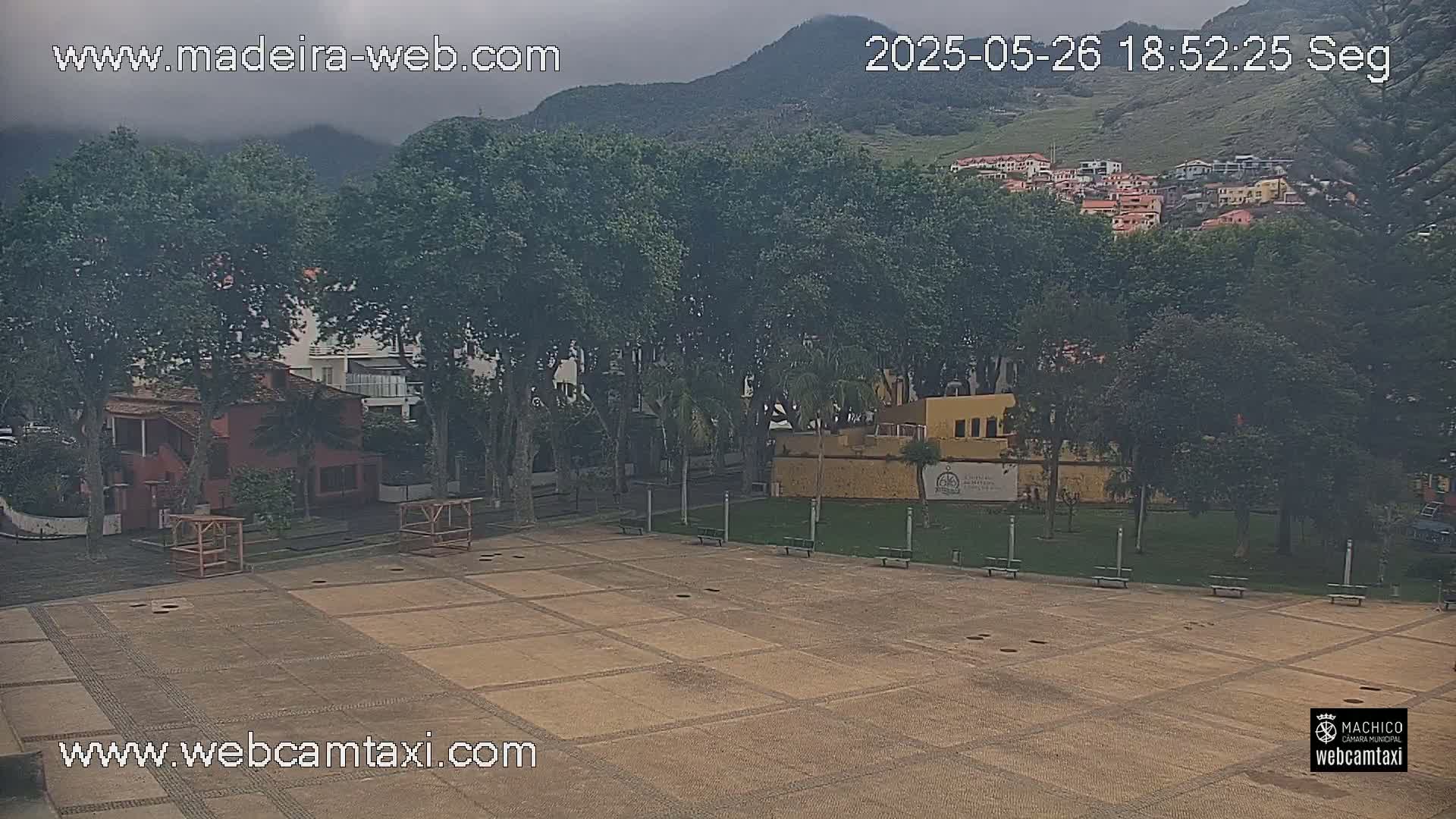 An overcast day reveals a paved plaza surrounded by leafy trees, buildings, and a grassy area, with hills visible in the background.