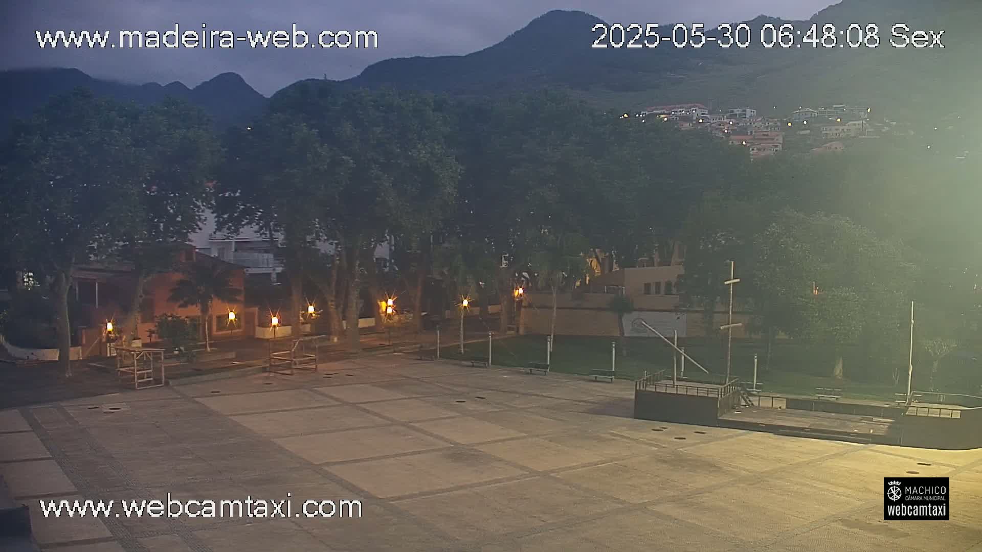 An empty plaza at twilight is surrounded by trees and buildings, with mountains in the background under a cloudy sky.