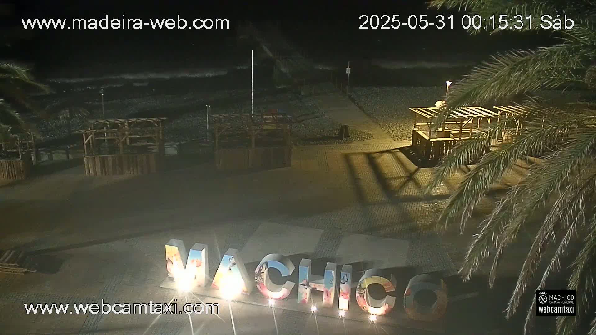 A nighttime beach scene shows dark water with waves, a pebble shoreline, several empty wooden structures, and large illuminated letters spelling out a place name.