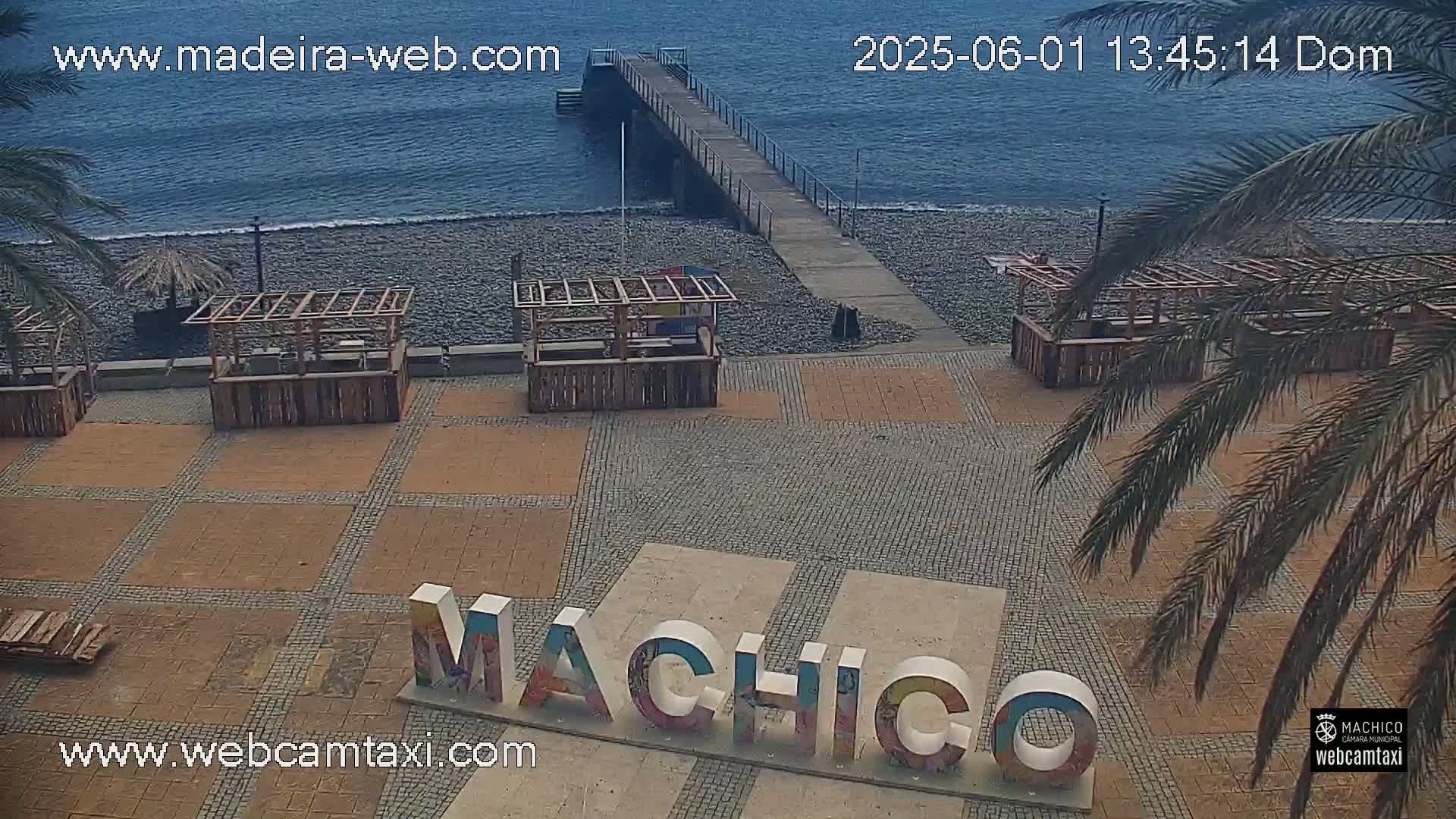 A beachside plaza with several wooden structures and a pier extending into a calm ocean, under a mostly clear sky.