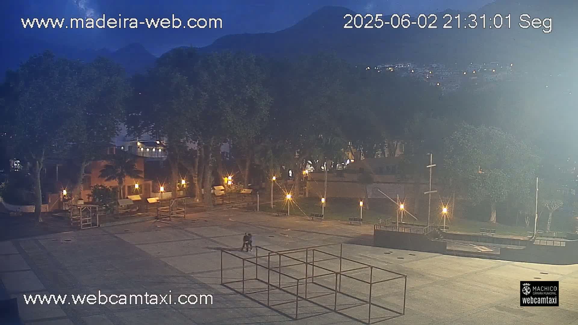 A nighttime, outdoor view of a town square with low light and a few people walking near a metal structure, surrounded by trees and buildings under a partly cloudy night sky with mountains in the background.