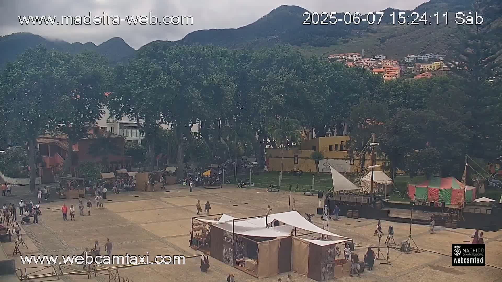 A town square with several market stalls and a replica ship, surrounded by trees and buildings under an overcast sky.