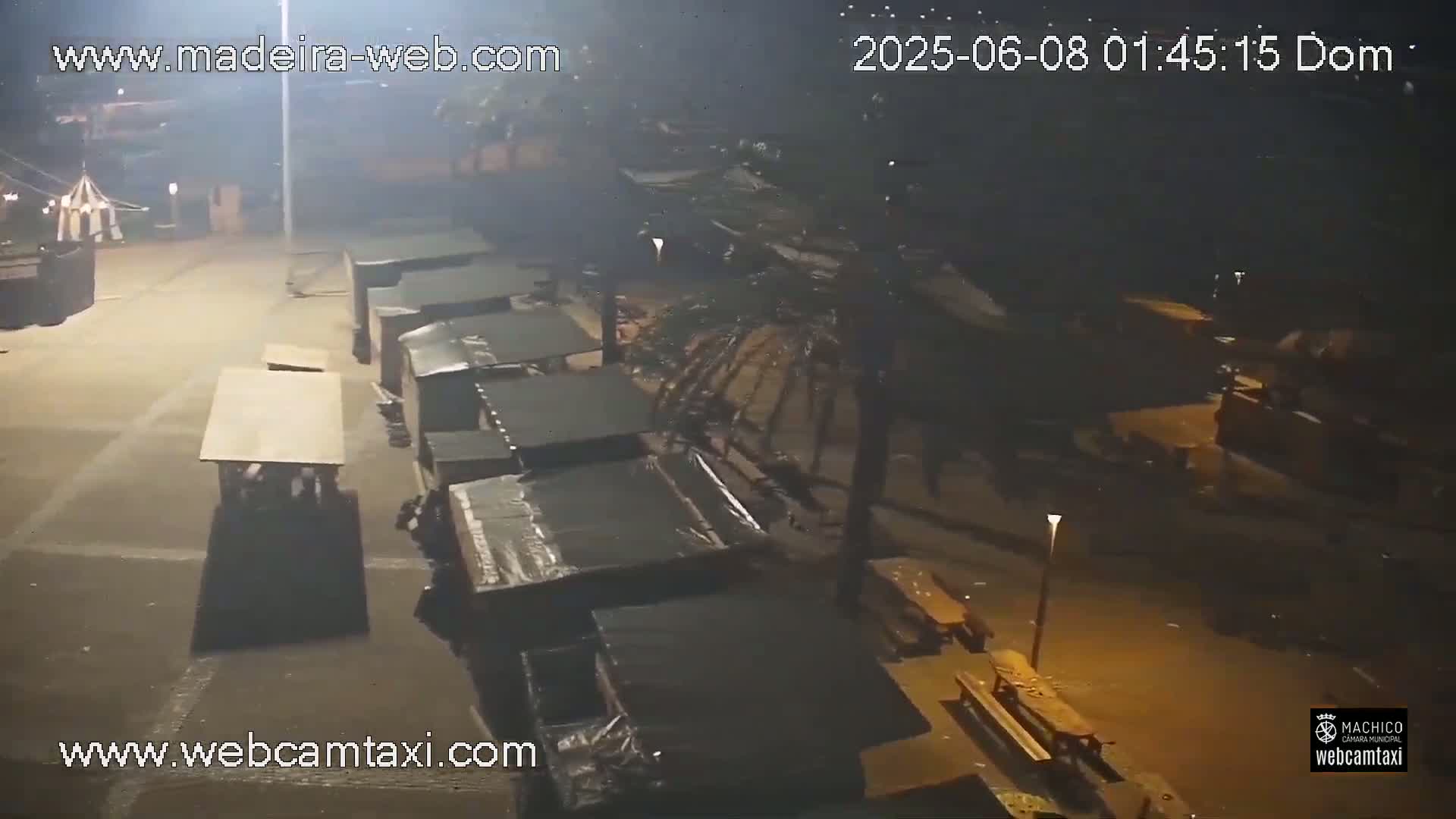 A nighttime, dimly lit outdoor area shows several dark stalls and picnic tables scattered across a paved surface.