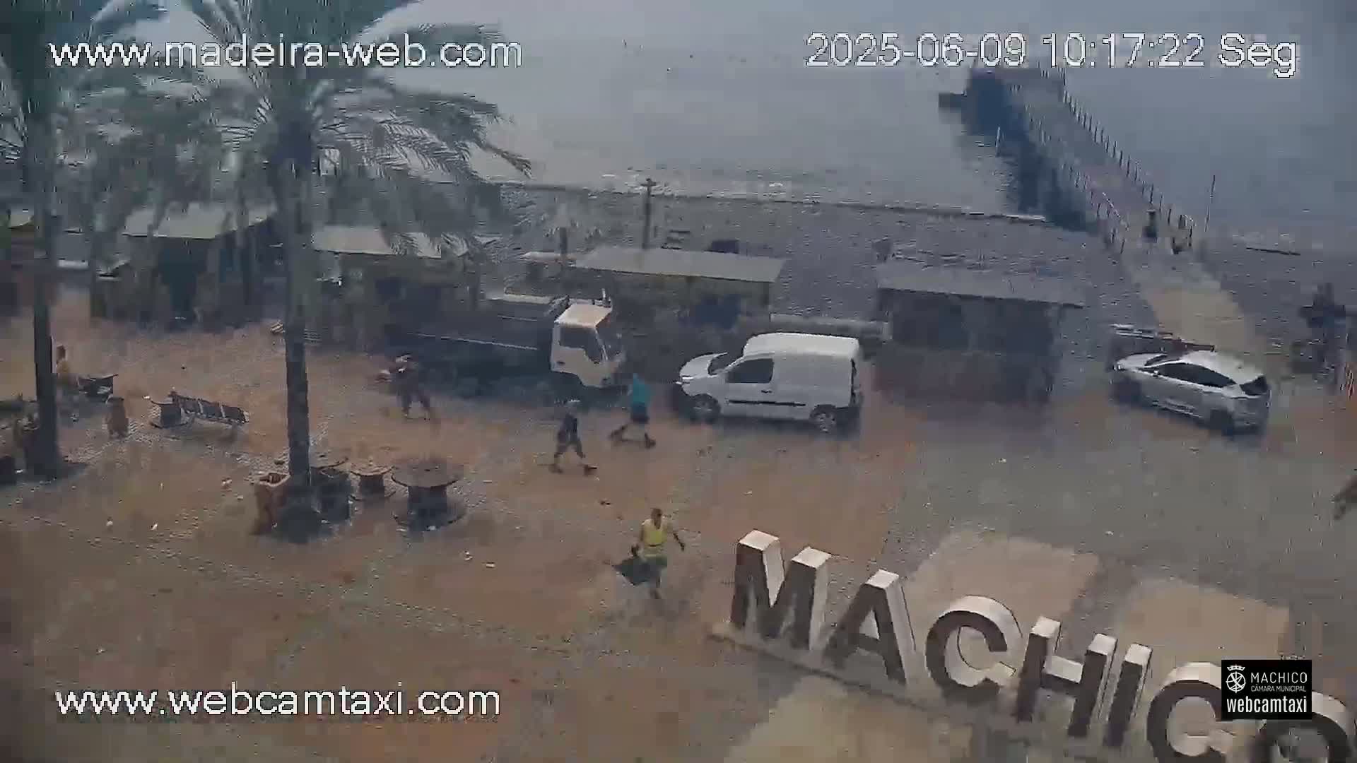 An overcast day shows a seaside area with several people walking near buildings, vehicles, and a large sign that reads "MACHICO".