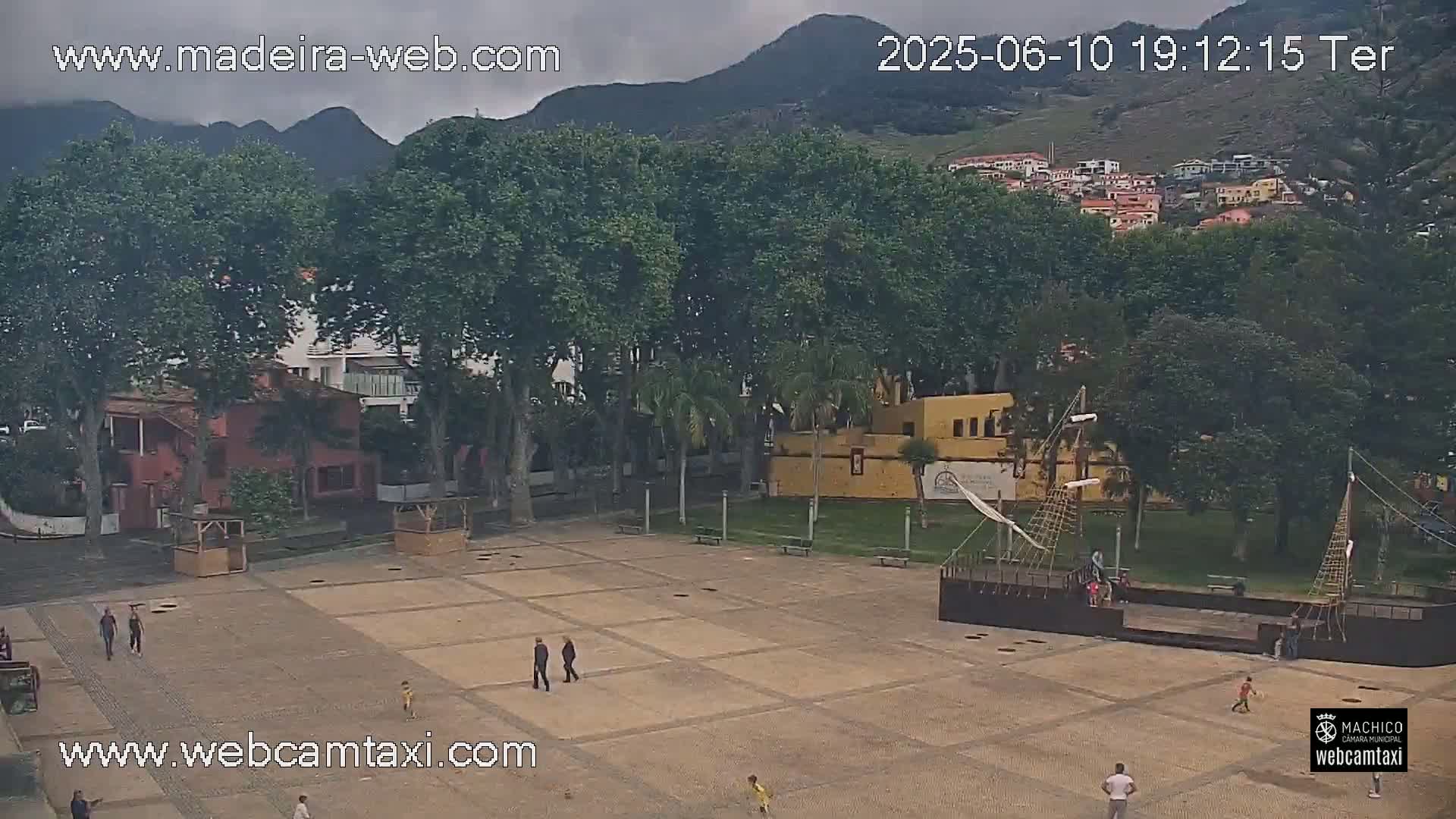 A mostly empty paved plaza with a few people, some trees, a building, and a large model ship, all under an overcast sky.