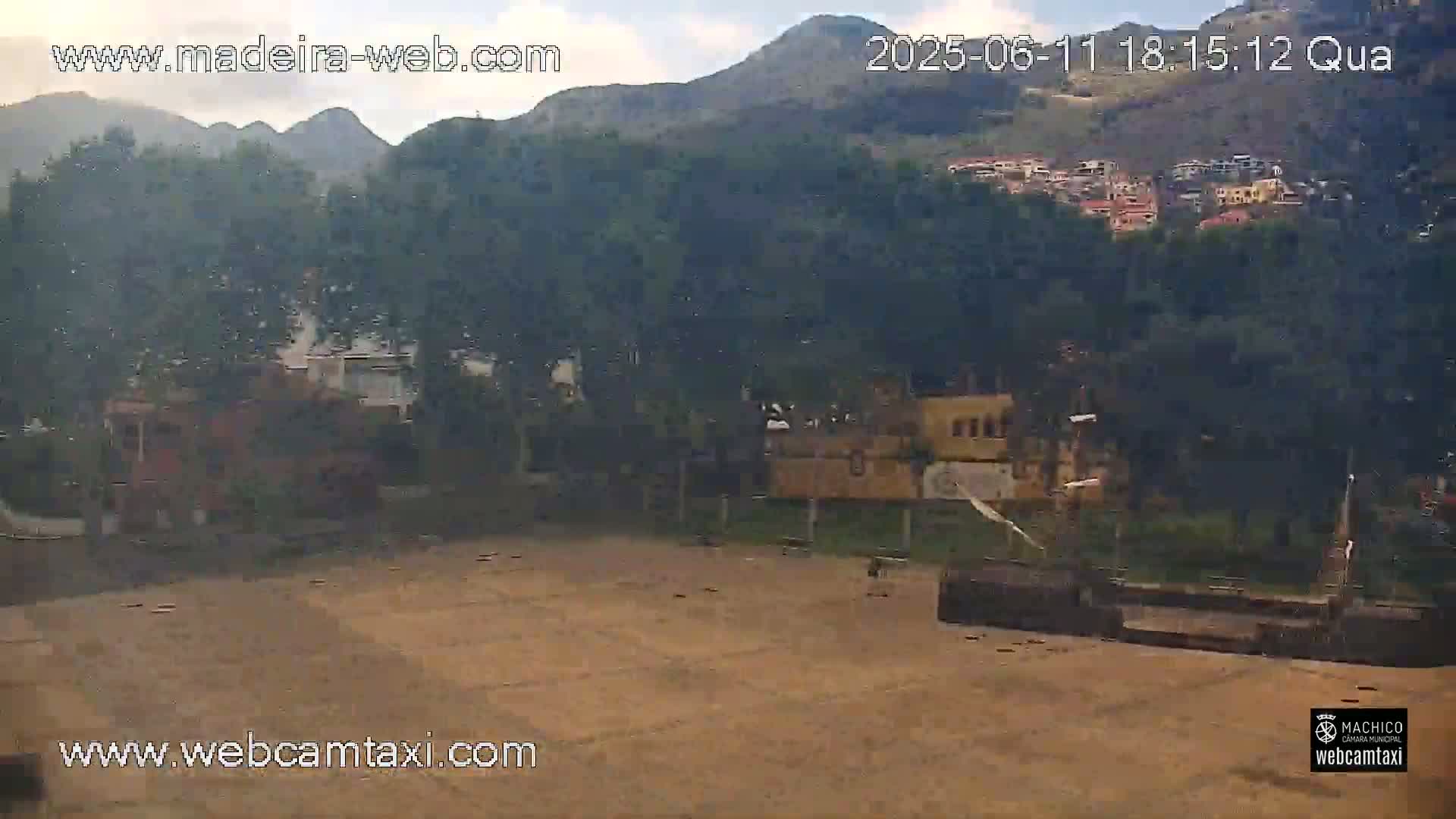 A mostly clear day reveals a tan, empty paved area, surrounded by greenery, buildings, and hills in the background.