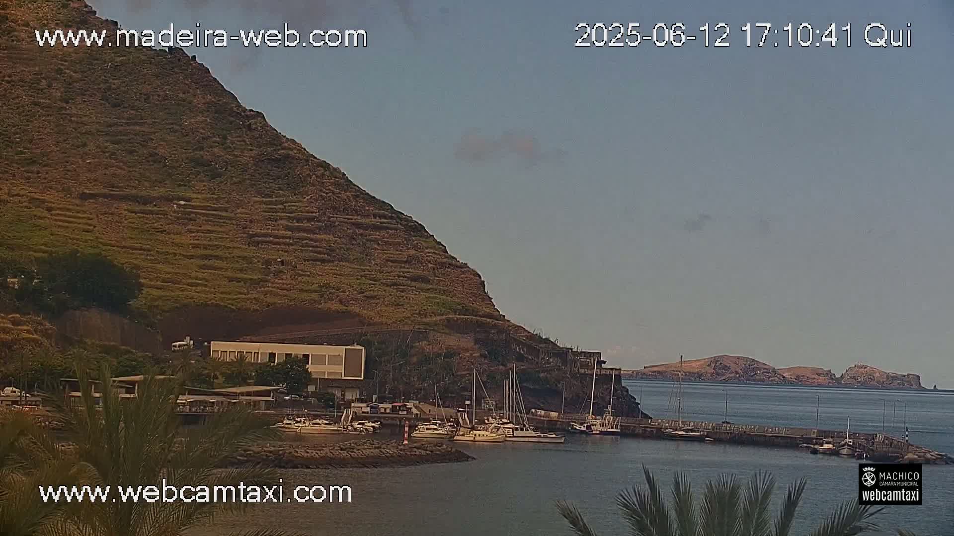 A small harbor with several boats is nestled below a steep, terraced hillside under a mostly clear sky.