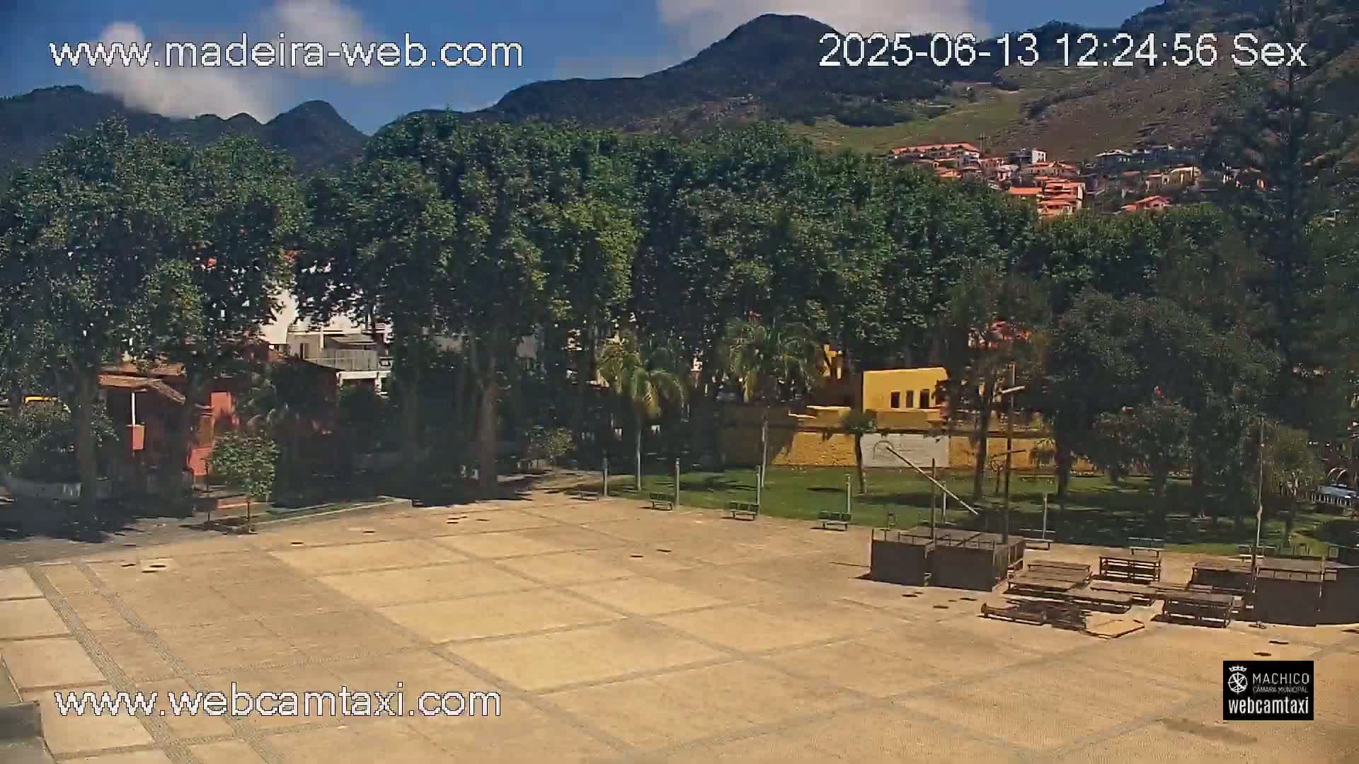 A paved plaza with park benches and a stage-like structure is surrounded by lush green trees and buildings, with mountains in the background under a mostly sunny sky.