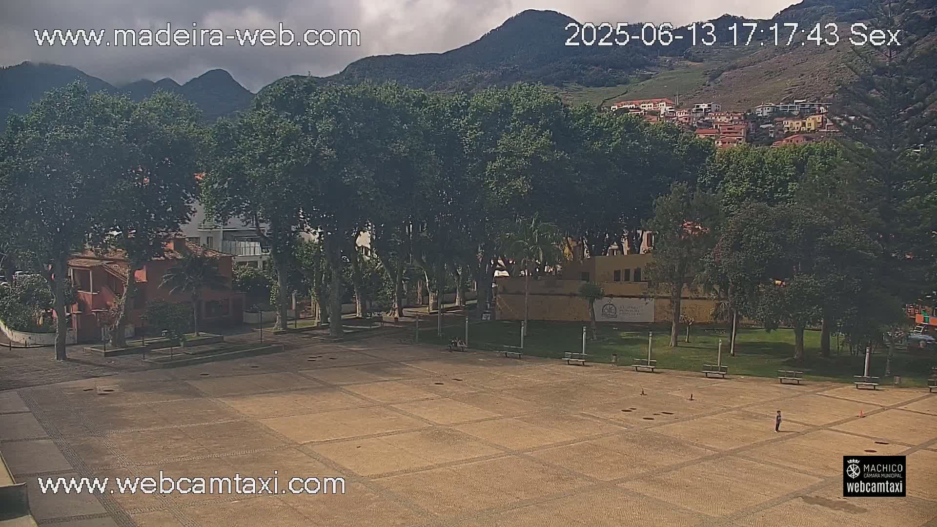 A mostly empty paved plaza is surrounded by lush green trees, buildings, and hills under a partly cloudy sky.