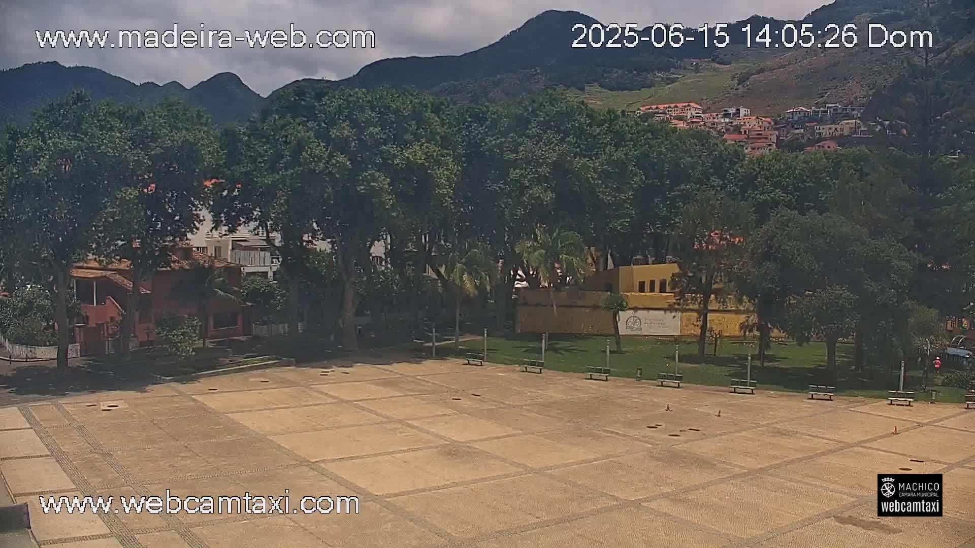 A paved plaza with benches sits in front of a row of trees and buildings, with mountains in the background under partly cloudy skies.