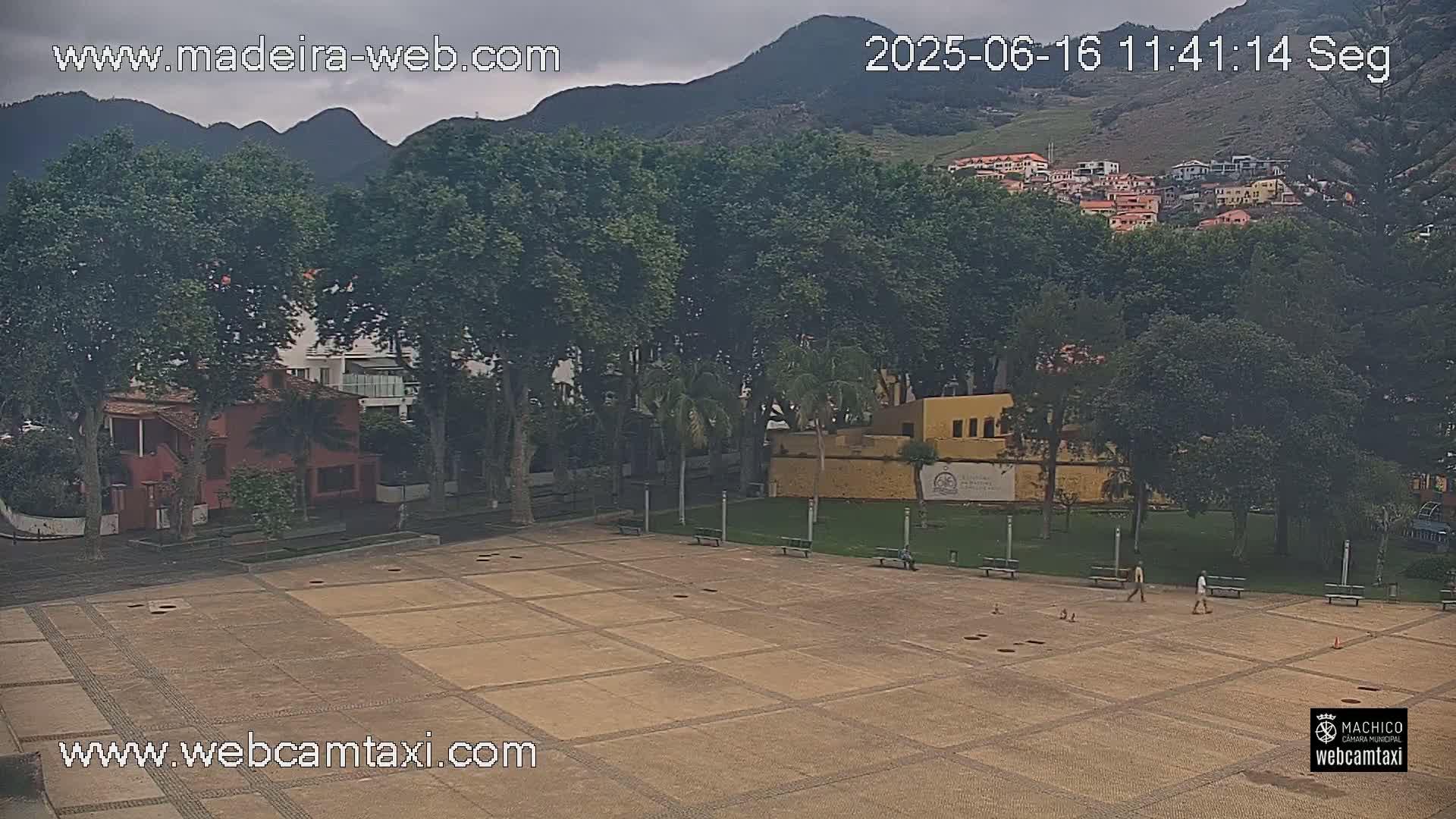 A mostly empty, paved plaza is surrounded by trees and buildings, with a few people walking and mountains in the background under an overcast sky.