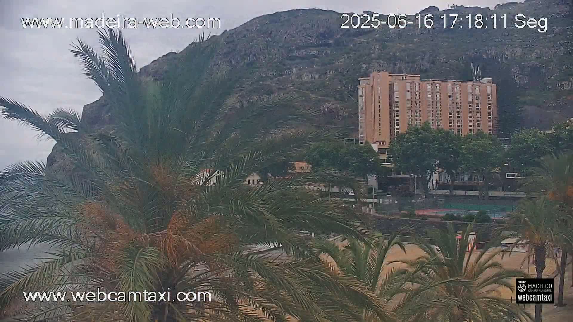A large, tan hotel is visible behind palm trees on a beach, with a mountain rising behind it all under an overcast sky.