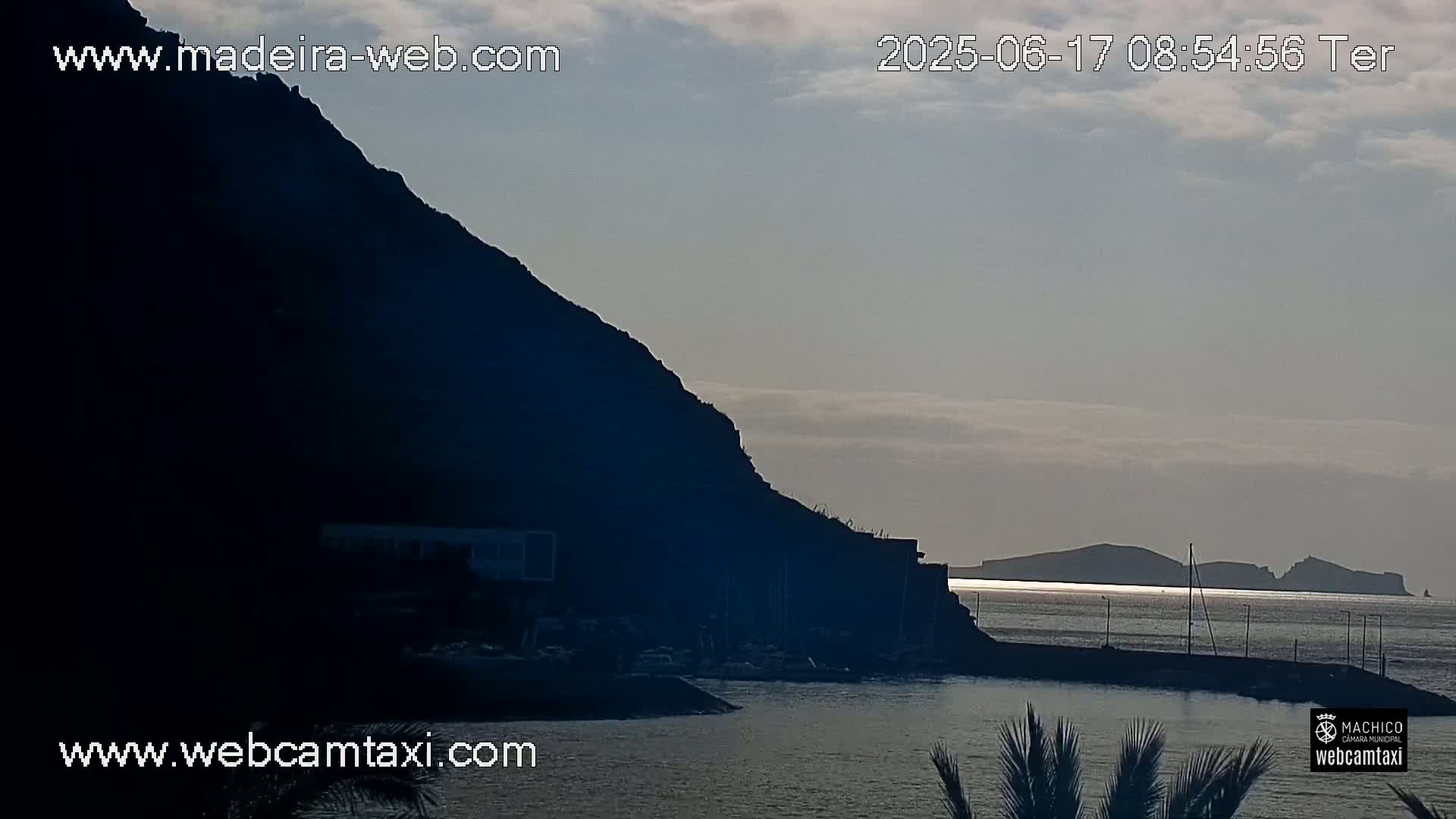 A dark, silhouetted cliff overlooks a calm body of water with distant islands under a mostly cloudy sky.