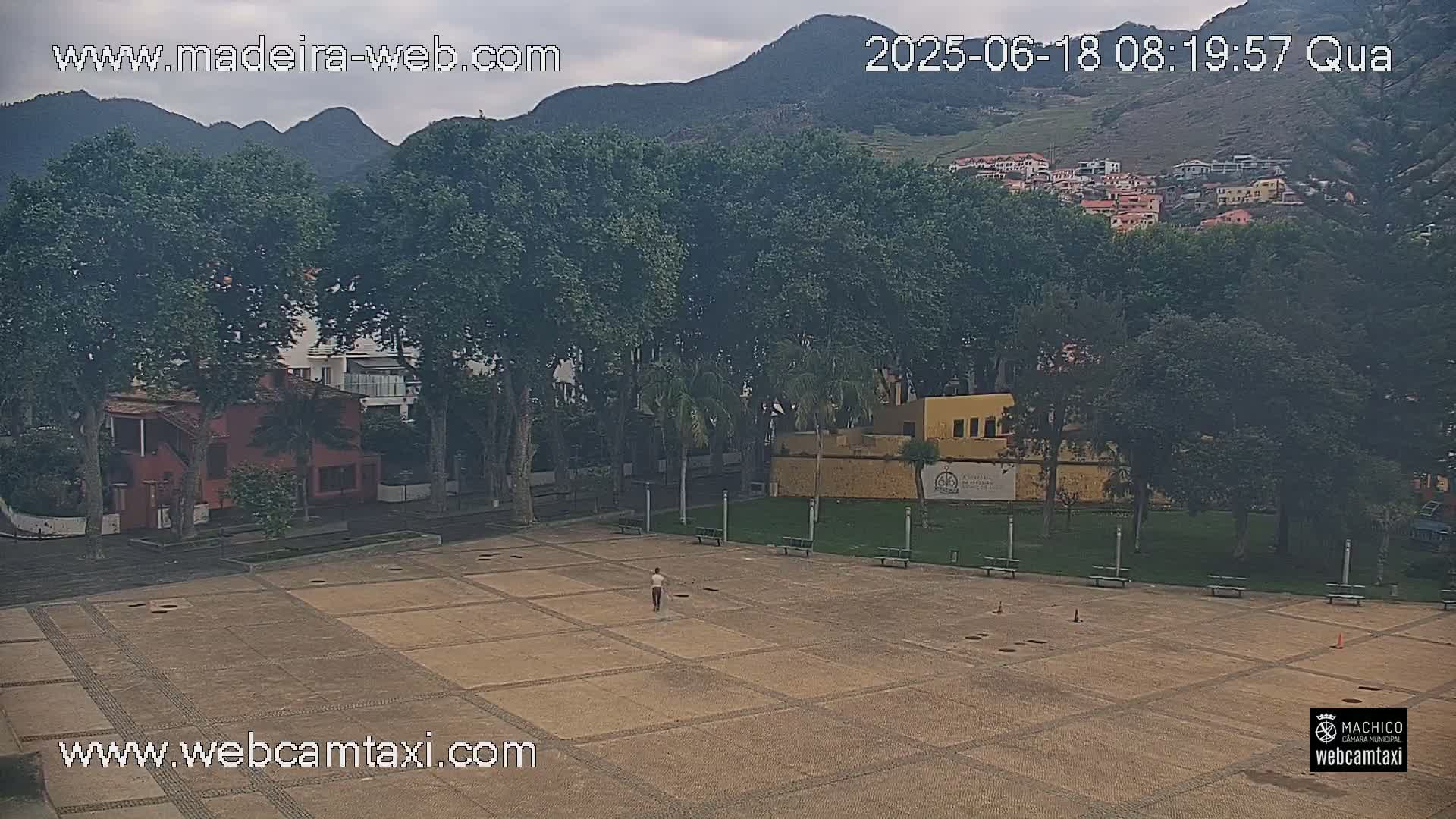 A person walks across a large, paved plaza surrounded by trees, buildings, and mountains under an overcast sky.