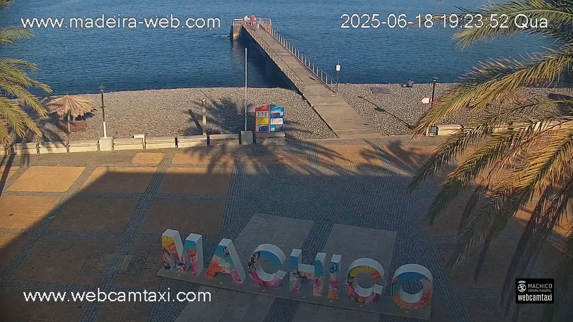 A paved plaza with a large "Machico" sign, palm trees, a pebble beach, and a pier extending into a calm body of water.