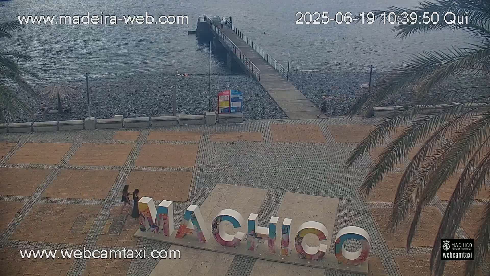 An overcast day reveals a pebble beach, a pier, a paved plaza with large "Machico" lettering, and palm trees, with a few people visible.