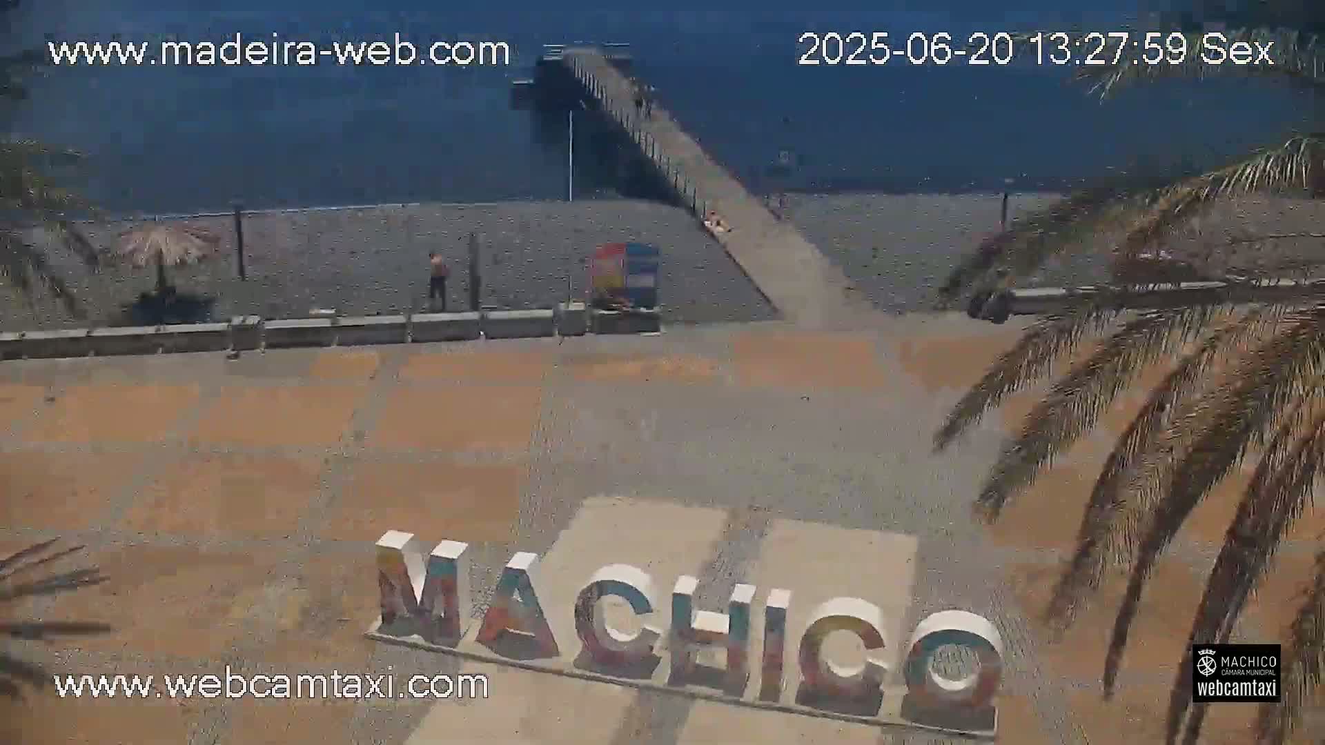 A beach scene on a sunny day shows a pier extending into dark blue water, a sparsely populated beach, and a large sign spelling out "Machico."