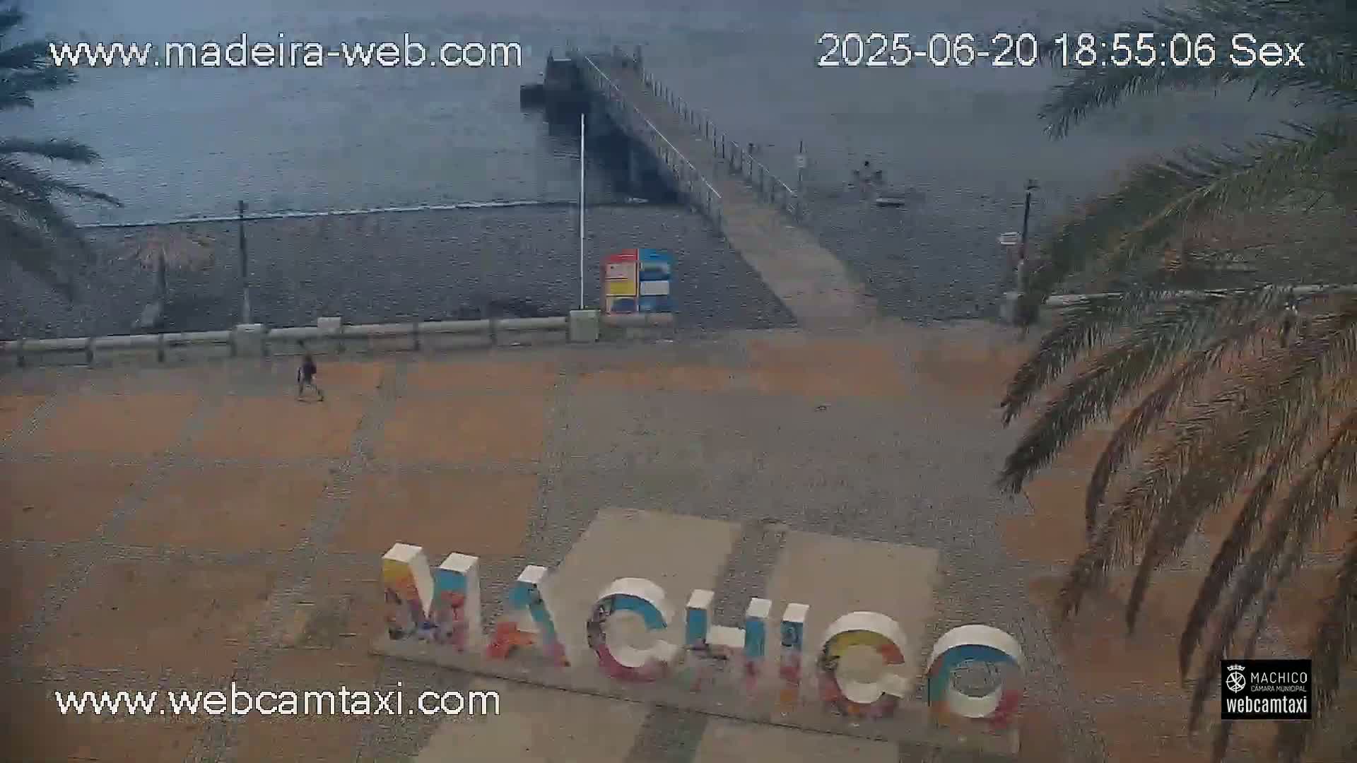 A person walks near a pier and beach, with large colorful letters spelling out a place name on a plaza; the weather is overcast.