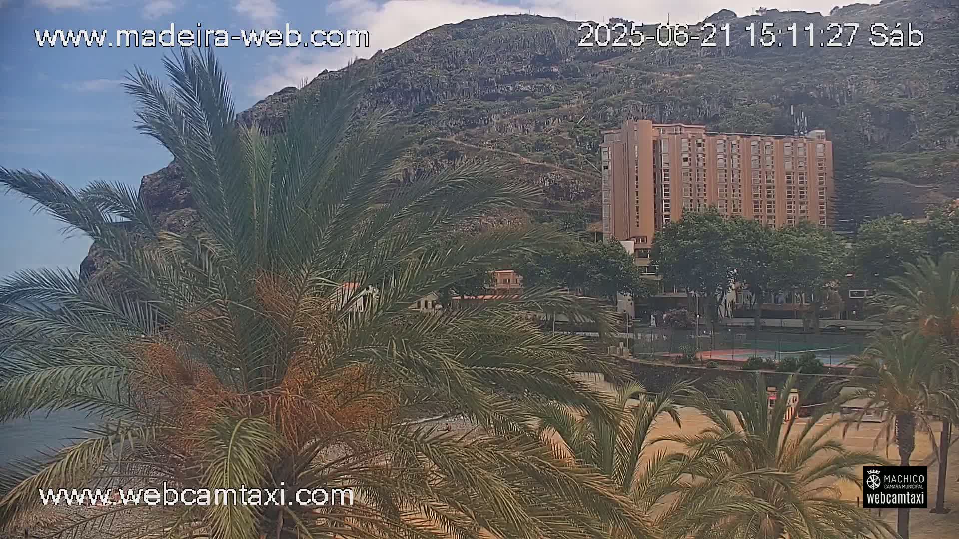 A partly cloudy day shows a beach, palm trees, a tall building, and a rocky hillside.
