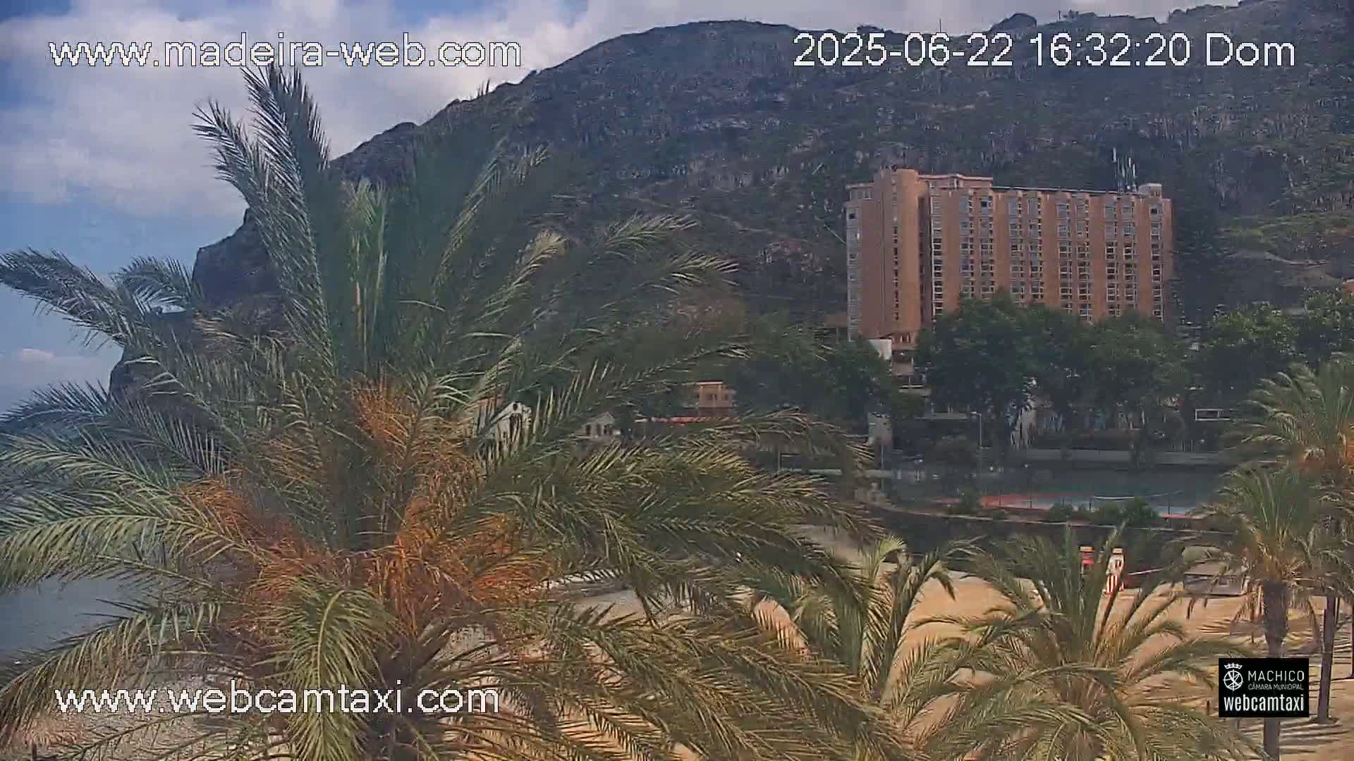 A large, tan hotel is partially visible behind palm trees, with a rocky mountain in the background under partly cloudy skies.