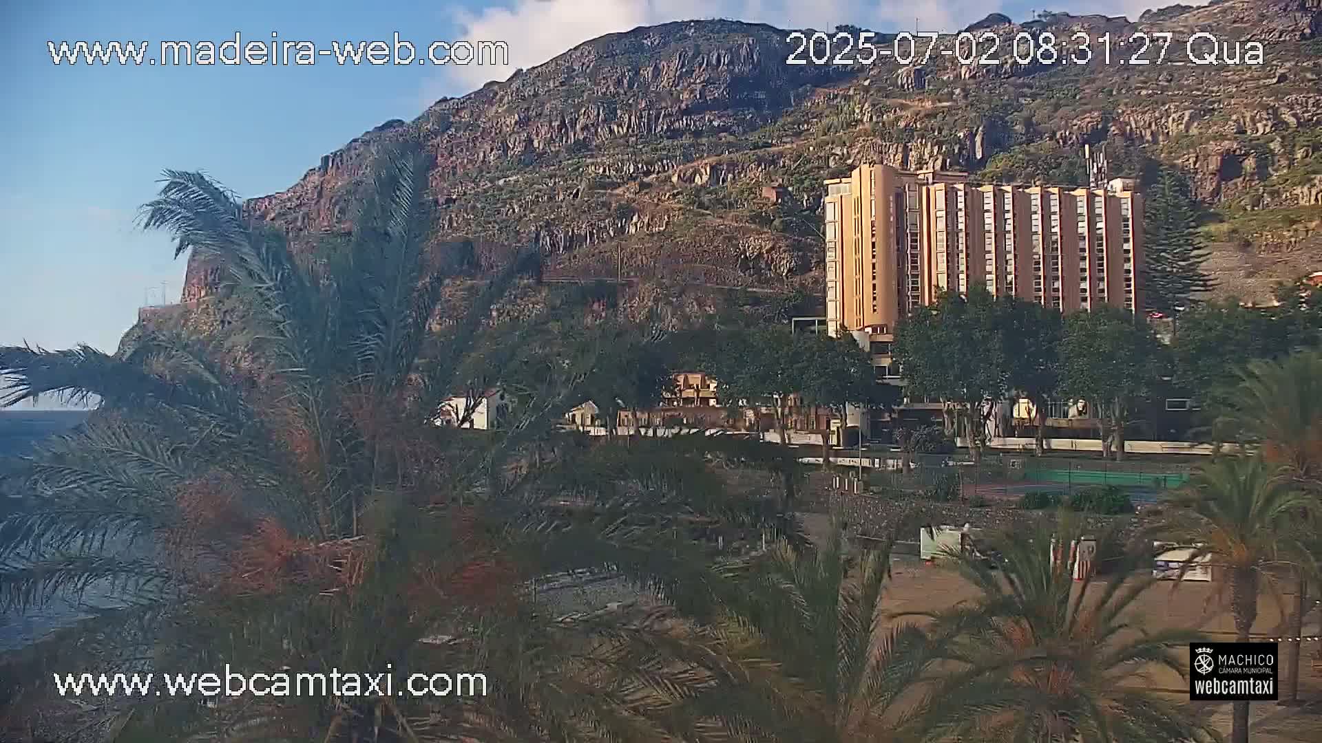 A multi-story building sits nestled amongst palm trees below a steep, rocky hillside on a partly sunny day.