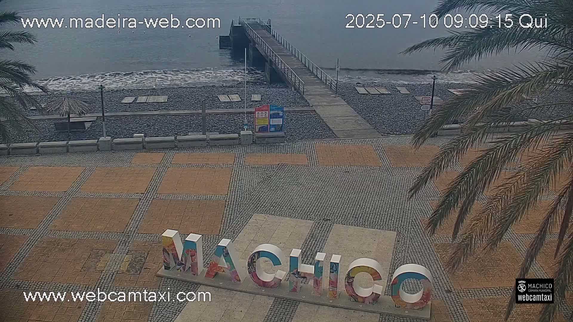 A pebble beach with a pier and calm water is viewed from above, showing a paved area with large letters spelling out a place name.