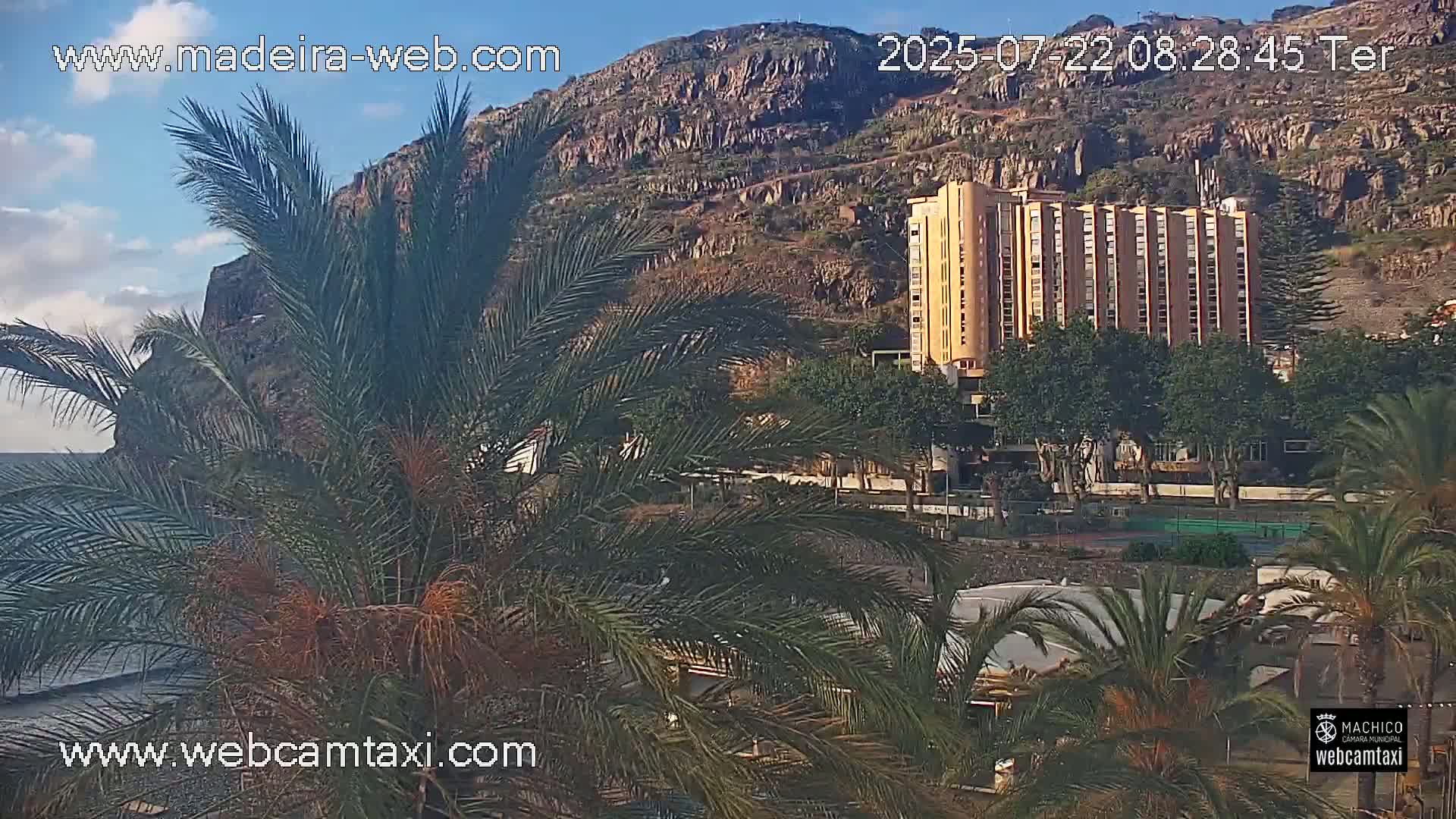 A multi-story building sits below a rocky hill overlooking a calm ocean, partially obscured by palm trees in a partly cloudy sky.