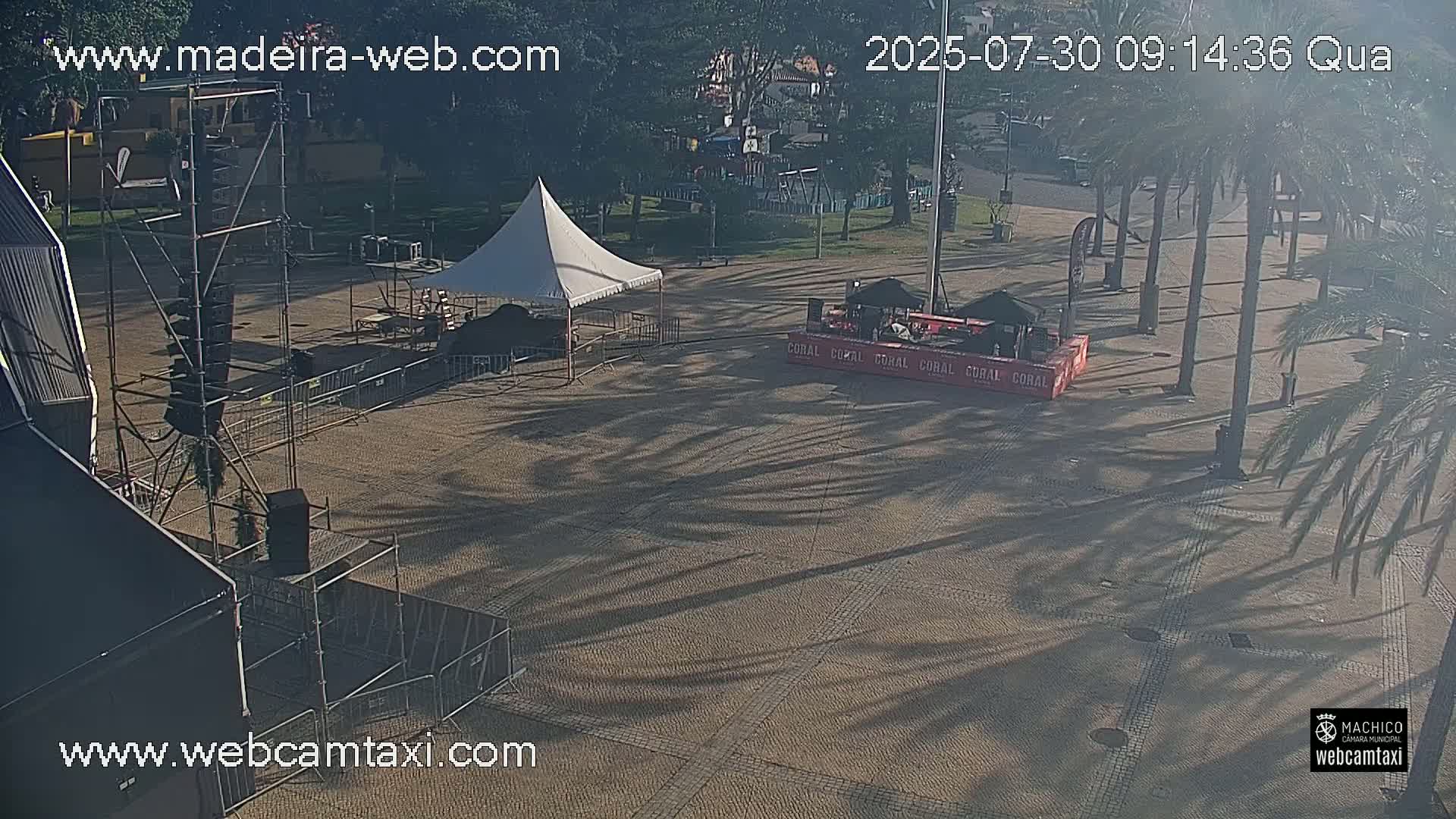 A paved plaza with palm trees casting long shadows in the morning sunlight, featuring a white tent and various sound and staging equipment.