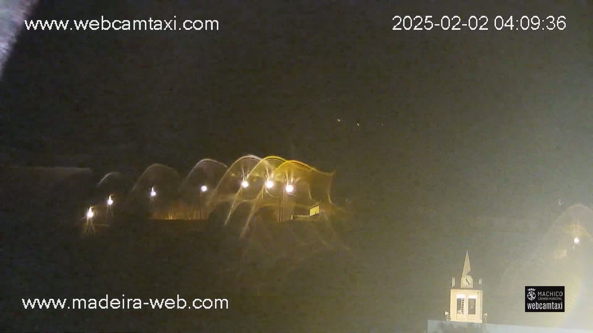 At night, illuminated water fountains arc over a dark landscape, with a small building visible in the lower right corner.