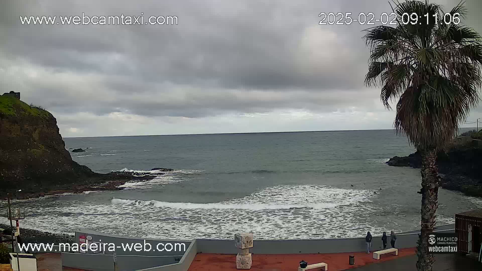 A partly cloudy day shows a rocky coastline with a calm ocean, small waves lapping the shore, and a few people standing near a palm tree.